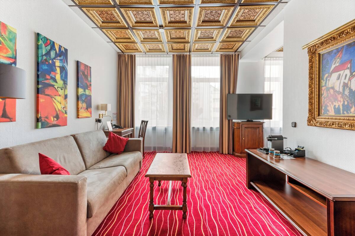 A hotel room with a red carpet, a beige sofa, a wooden table, a TV, and artwork on the walls.