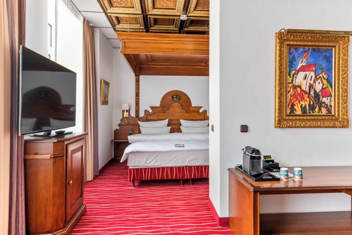 A hotel room with a wooden headboard, a television on a cabinet, a red carpeted floor, a desk with a coffee maker, and a painting on the wall.