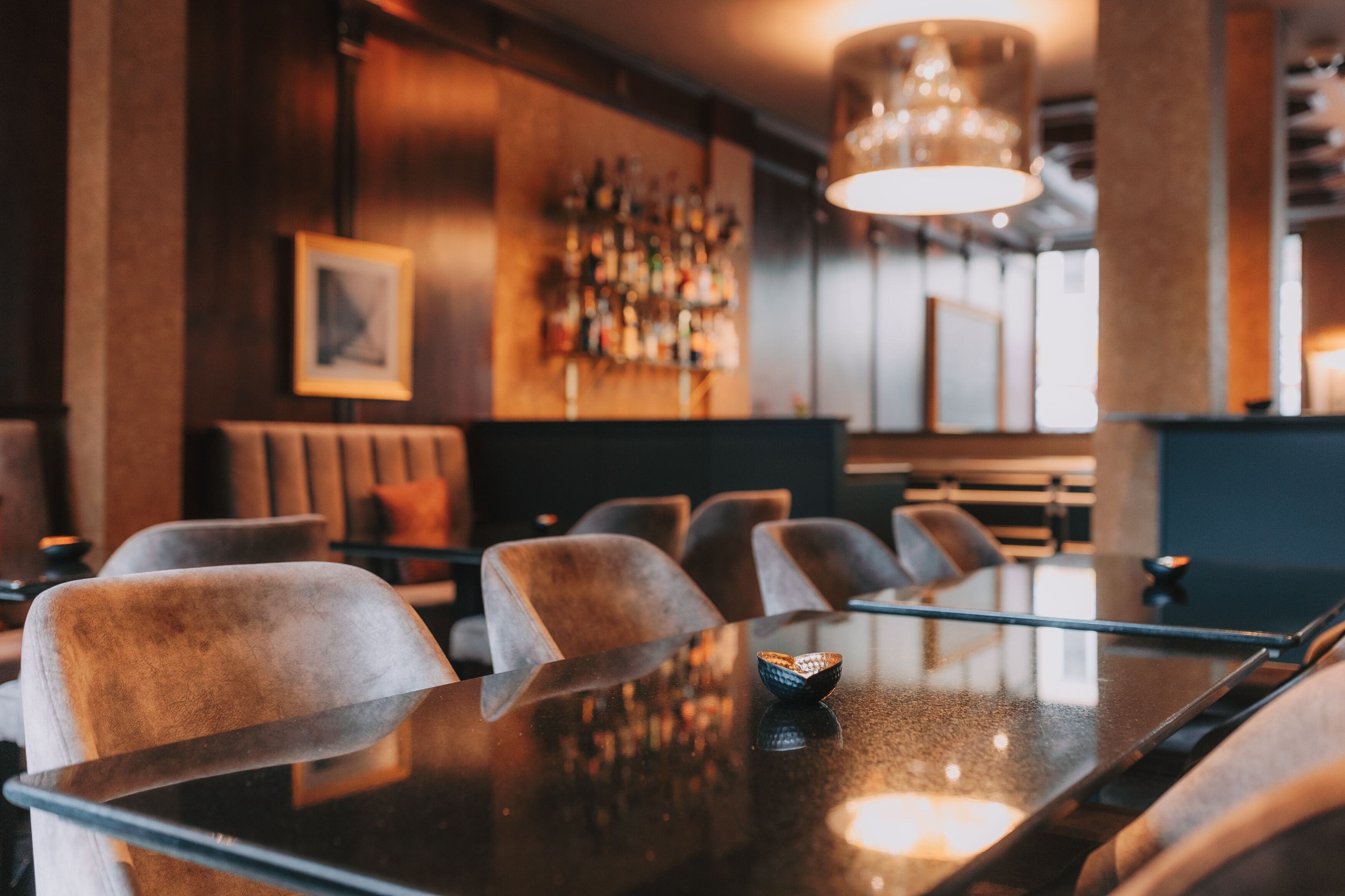 Zona de bar con una mesa larga, sillas acolchadas y una barra llena de botellas.
