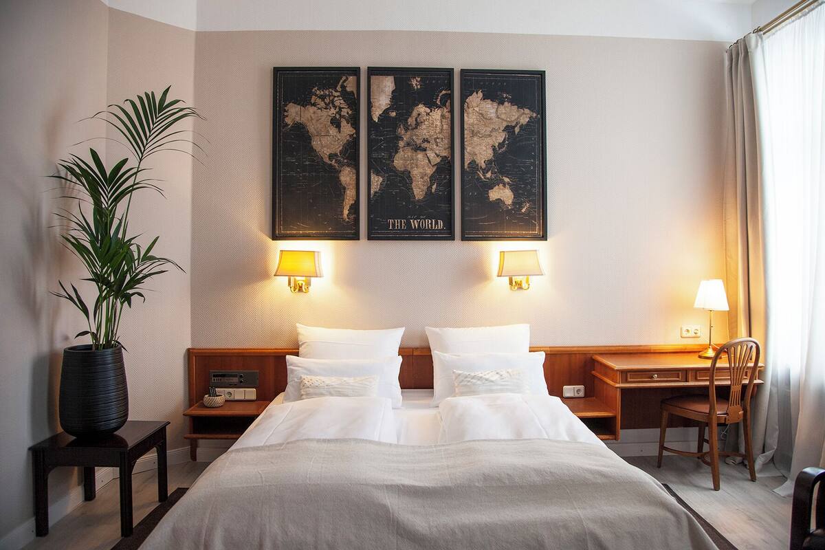 Una habitación de hotel con una cama grande, dos mesitas de noche, un escritorio, una silla, una planta en maceta y tres mapas del mundo en la pared.