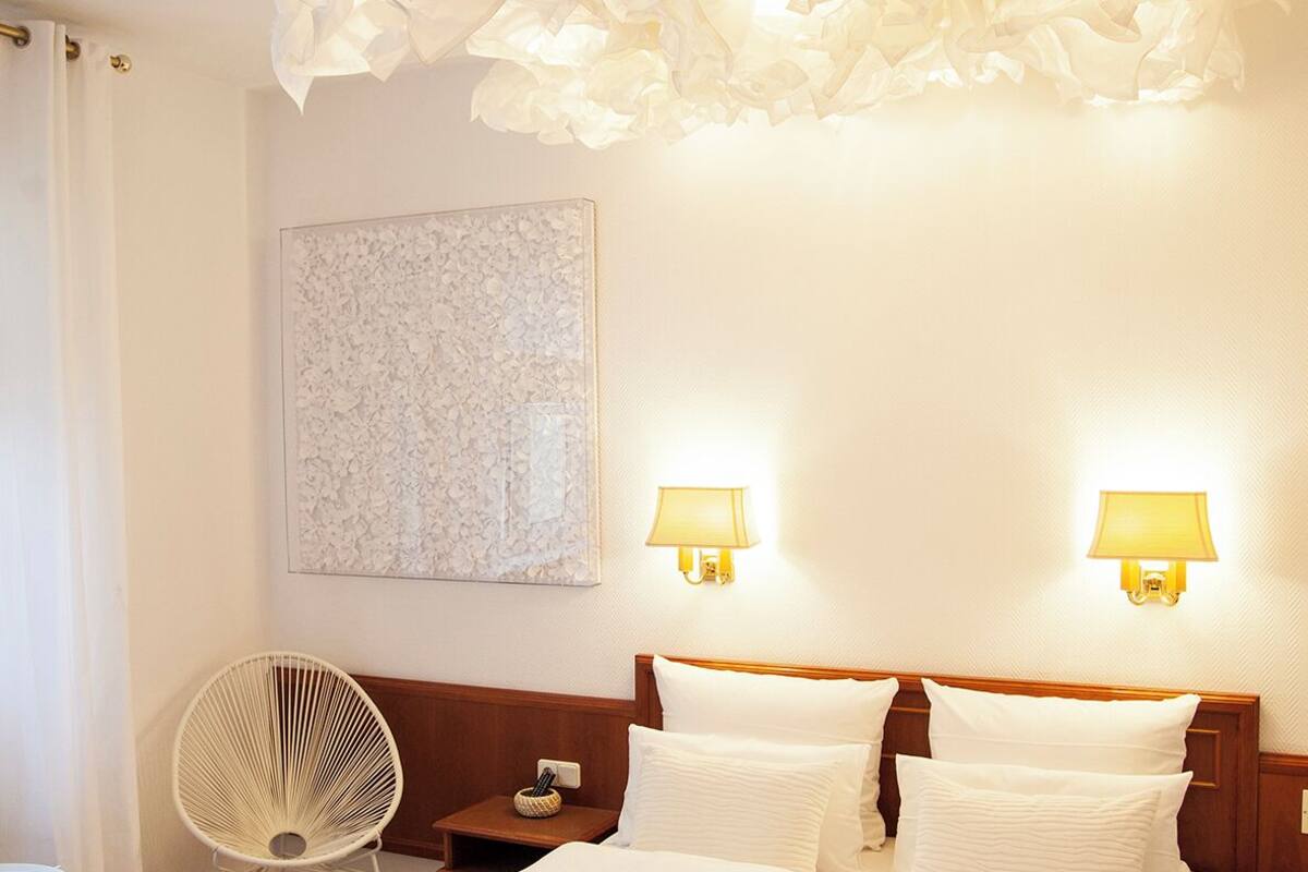 Una habitación de hotel con una cama grande, dos lámparas de noche, una obra de arte en la pared y una lámpara de techo original.