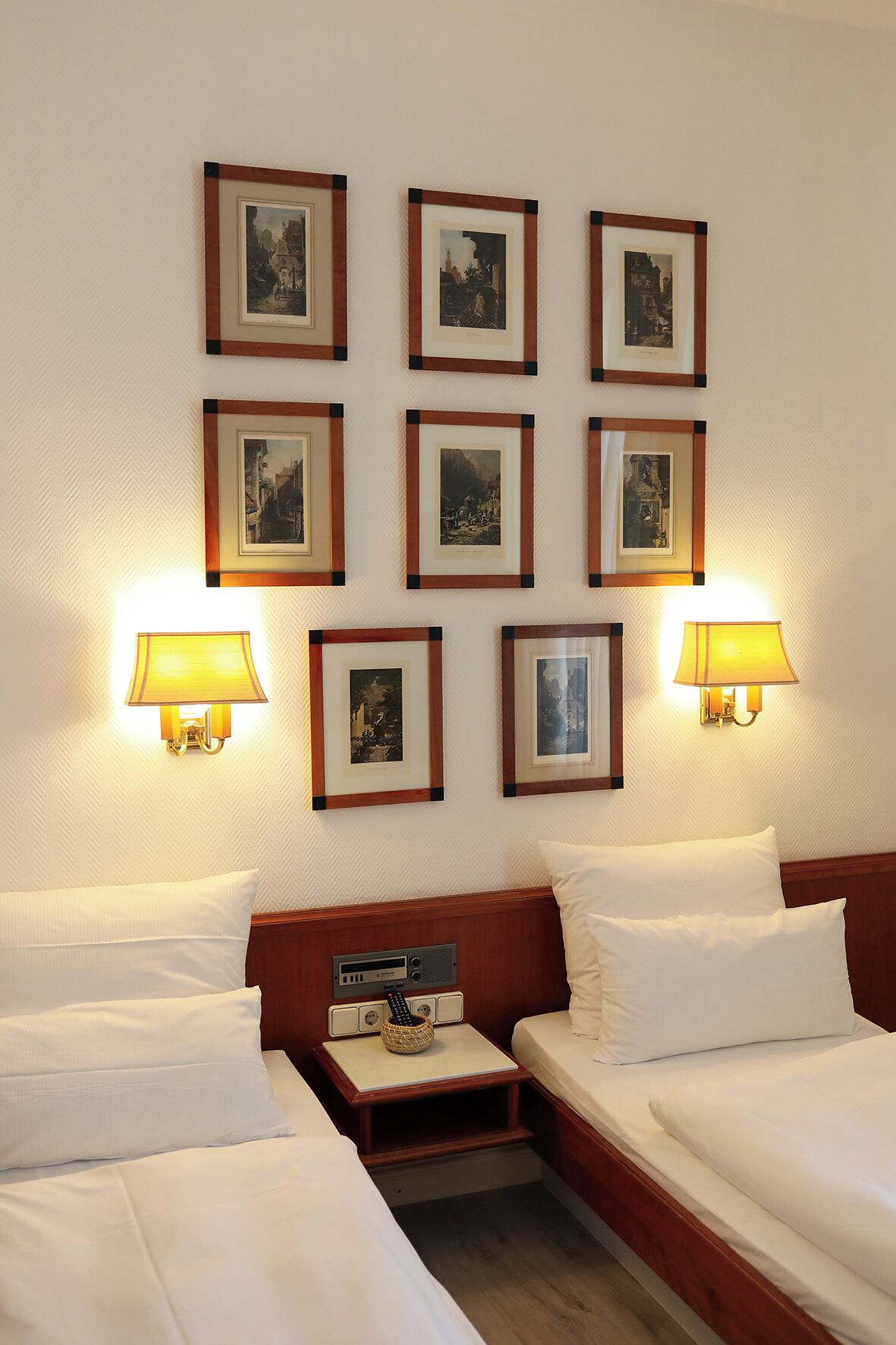 Habitación de hotel con dos camas, una mesita de noche, una lámpara y cuadros en la pared.