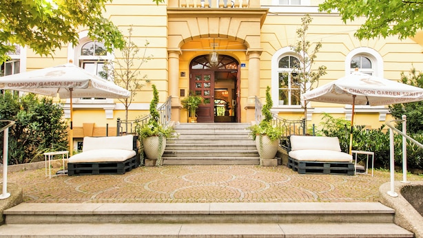 A hotel entrance with a paved courtyard, outdoor seating, and a staircase leading to the entrance. - Boutique Hotel Krone München (Munich)