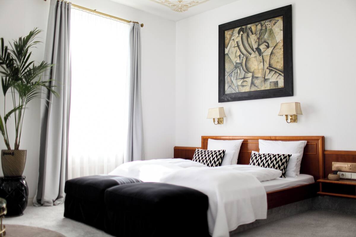 Habitación de hotel con cama, dos lámparas de noche, un cuadro abstracto grande, una planta en maceta y una ventana con cortinas.