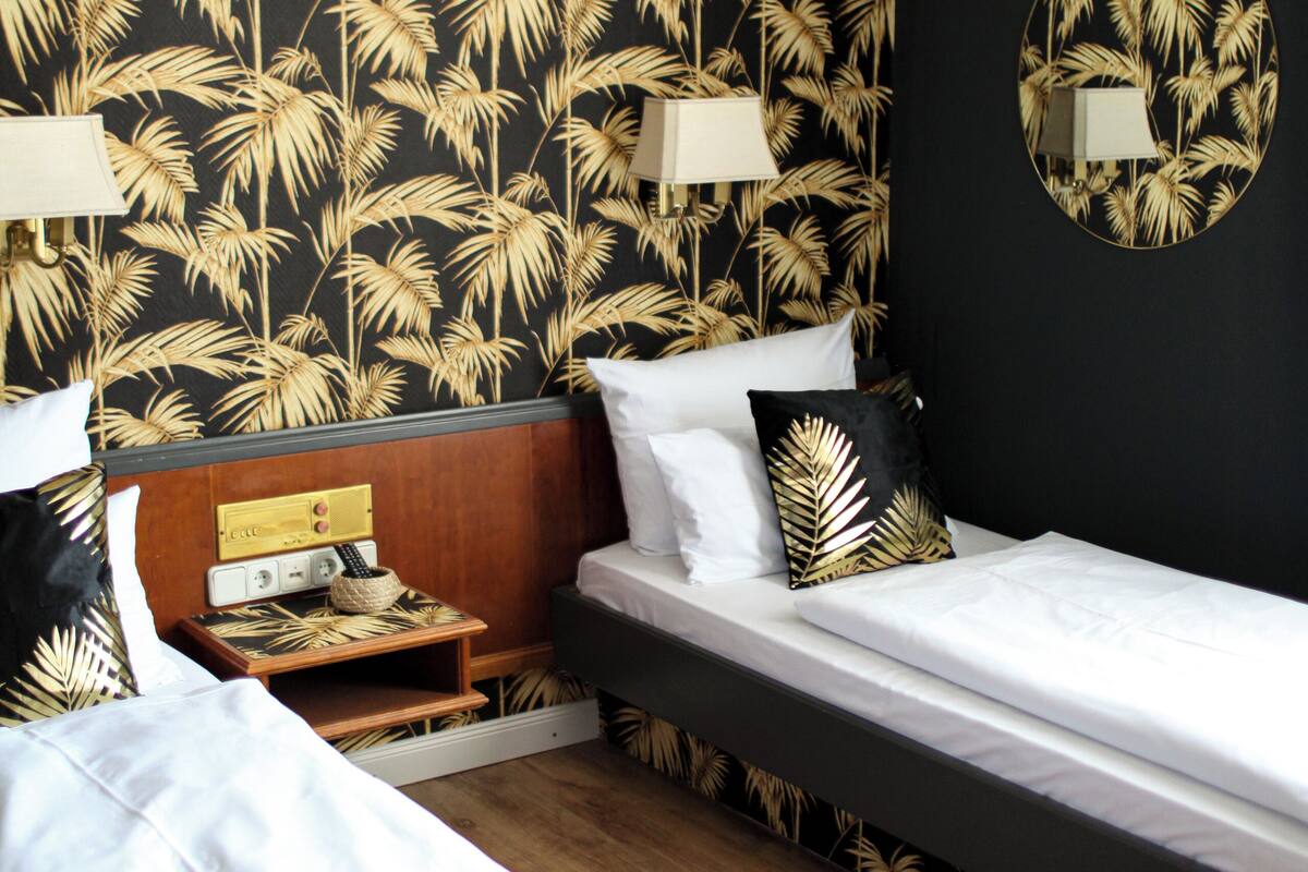 Habitación de hotel con dos camas, una mesita de noche de madera, un espejo y una pared con estampado de hojas de palmera.