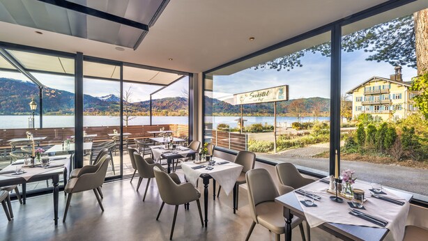 A restaurant with tables and chairs, large windows offering a view of a lake and mountains. - Seehotel zur Post (Tegernsee)