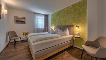 A hotel room with a large bed, two chairs, a small table with a vase of sunflowers, and a painting on the wall.