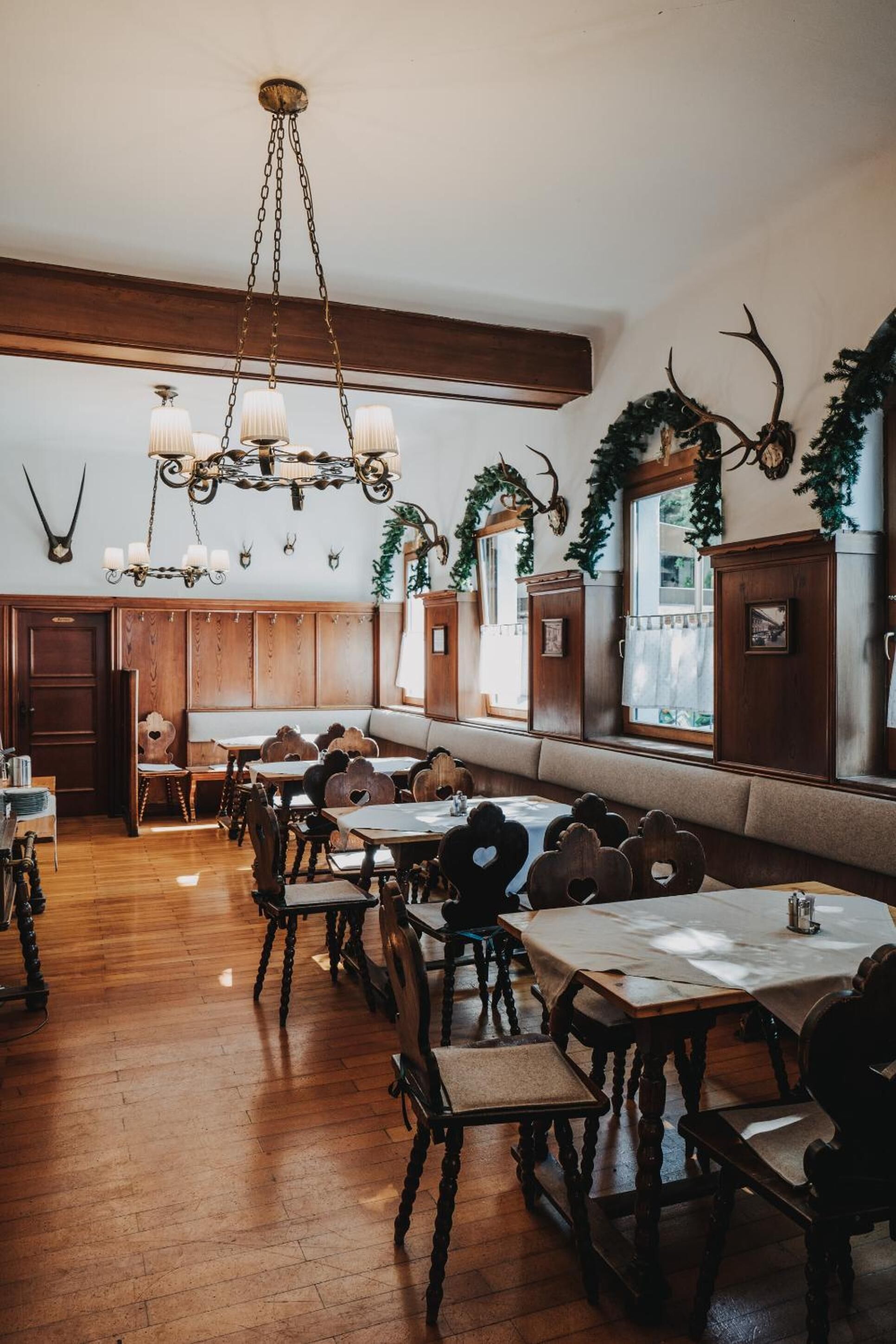 A rustic dining area with wooden tables and benches, a chandelier, antler decorations, and a view of the outdoors through large windows.