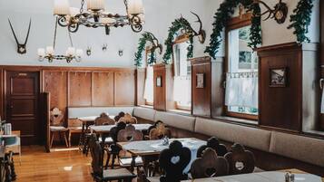 A rustic dining area with wooden tables and benches, a chandelier, antler decorations, and a view of the outdoors through large windows.