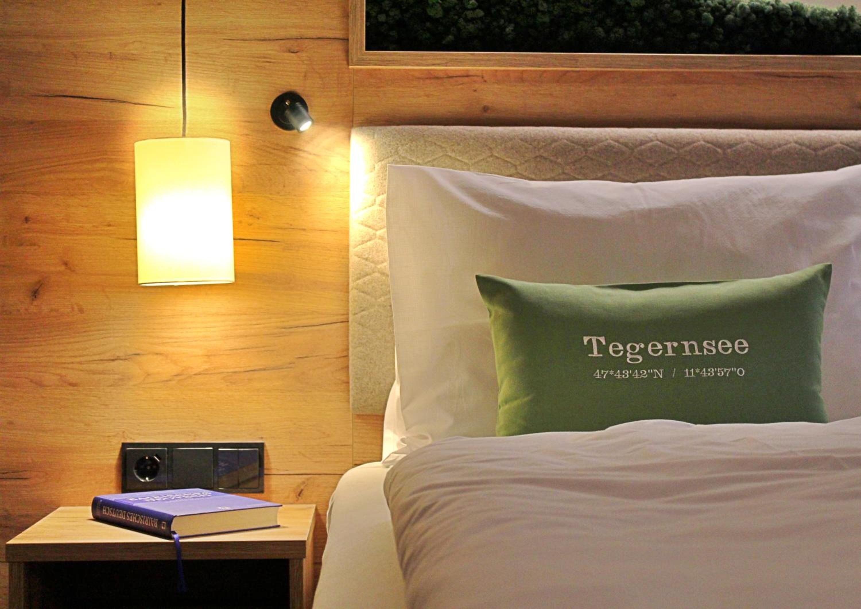 Una cama bien hecha con un almohadón verde que dice 'Tegernsee' y coordenadas, una mesita de noche con un libro, una lámpara y un altavoz.