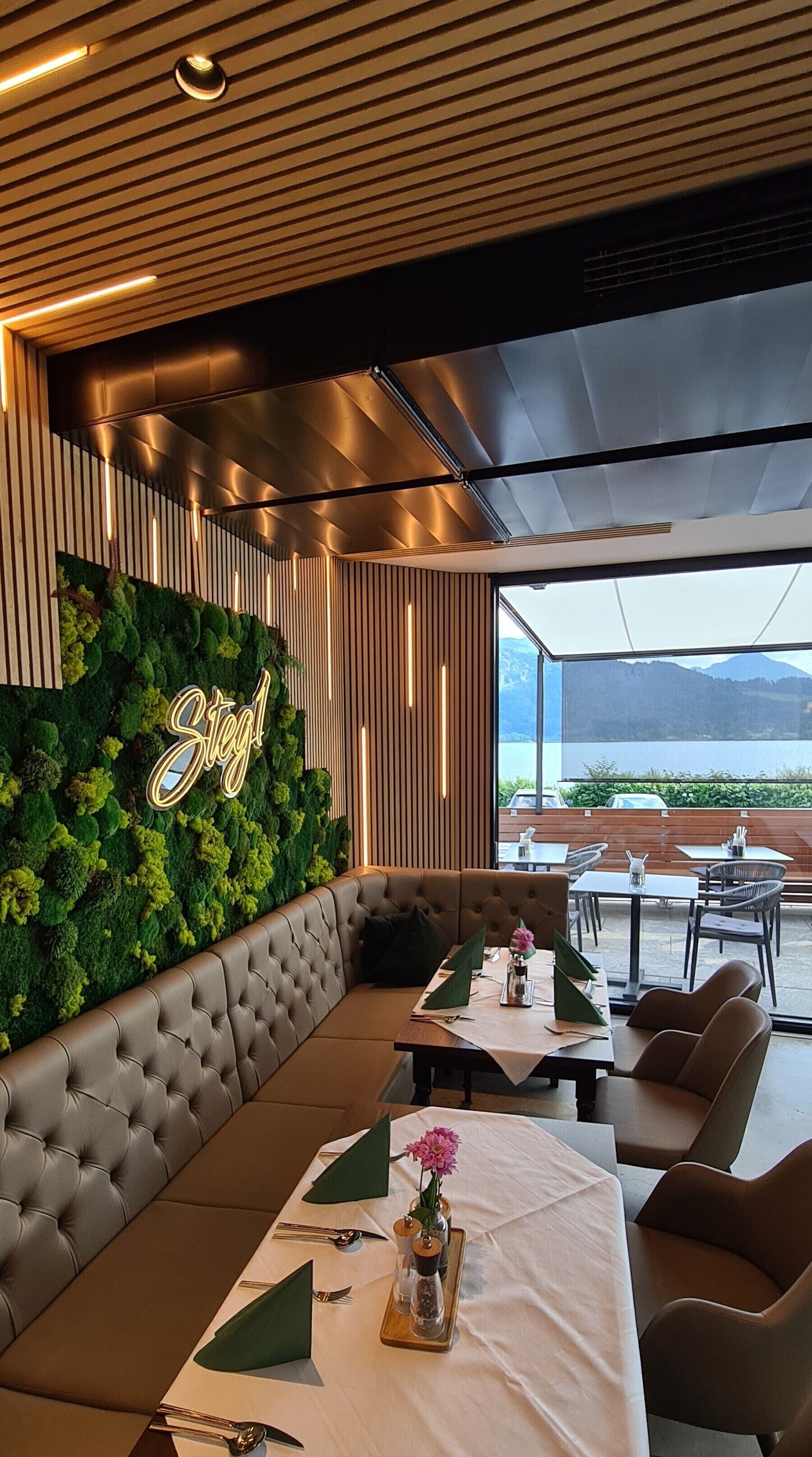 Un restaurante con una decoración moderna, que presenta una pared verde con el nombre 'Steg1' y vistas a un lago y montañas.