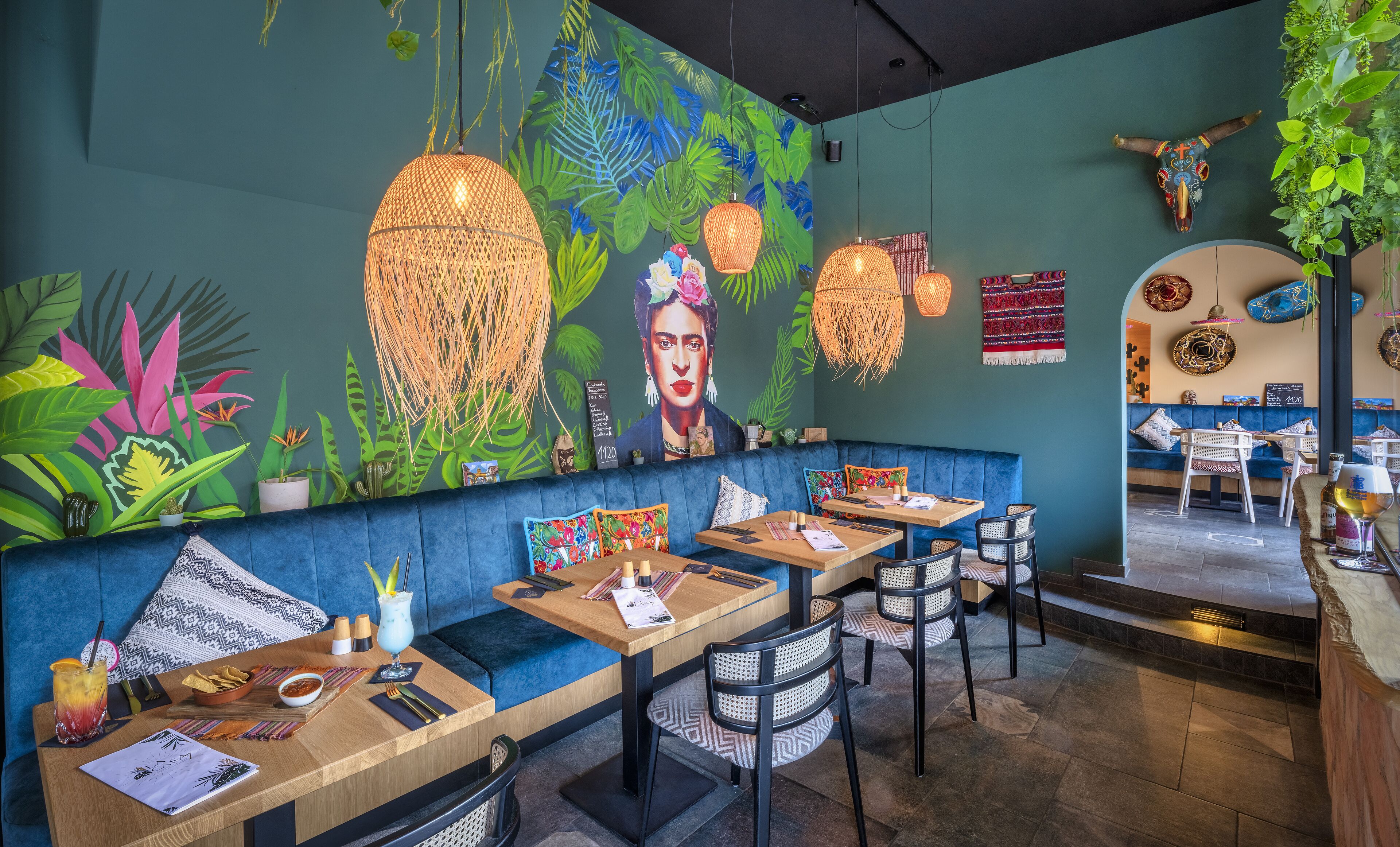 Un restaurant doté d’un coin salon avec des bancs rembourrés bleus, de luminaires suspendus en fibres tressées et d’une fresque représentant un feuillage tropical et le portrait d’une femme.