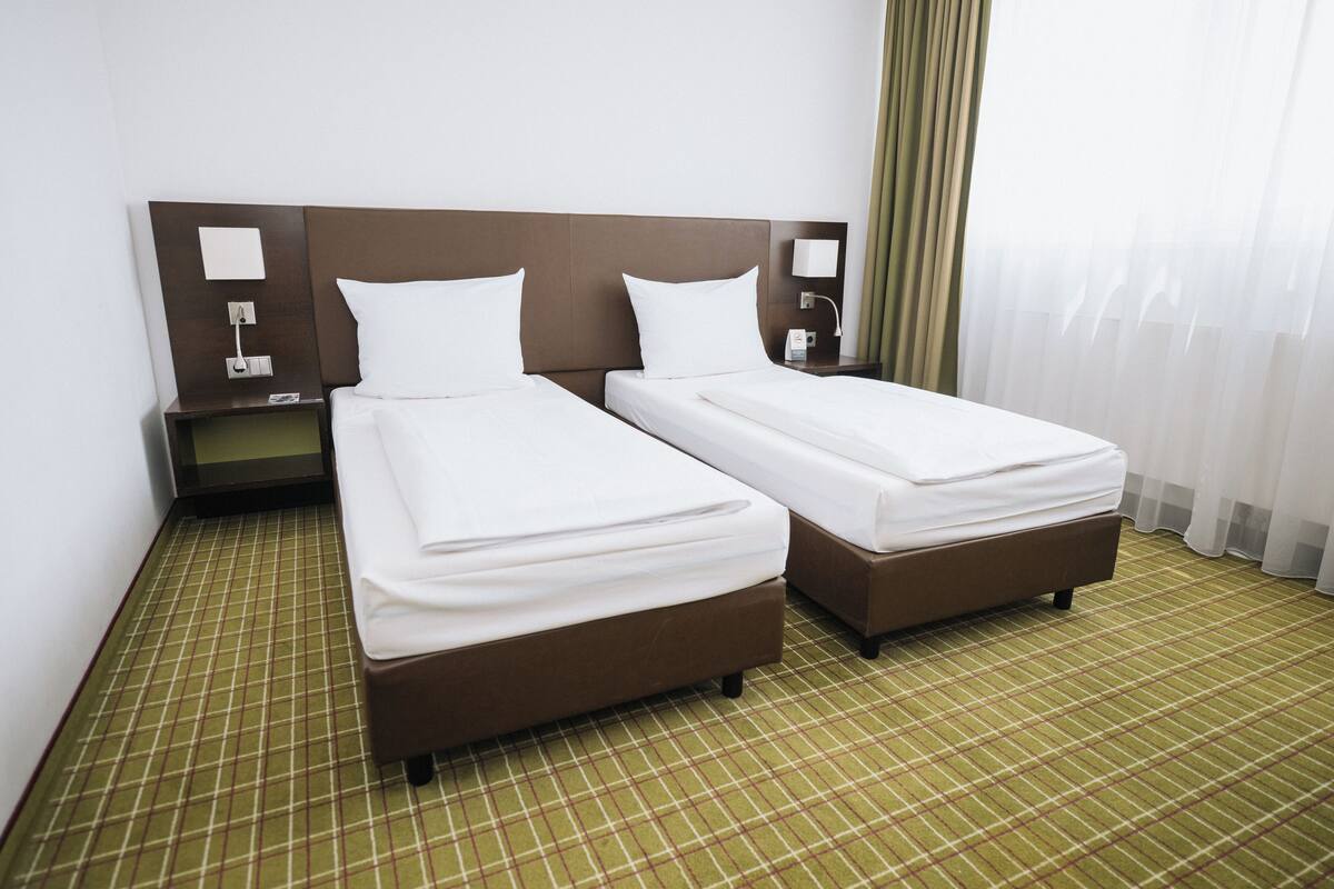A hotel room with two single beds, a nightstand with a lamp, and a check-in desk.