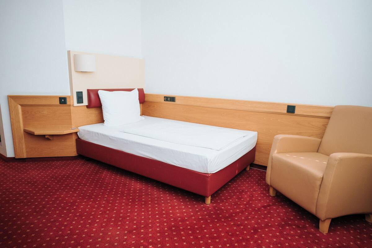 A hotel room with a single bed, a wooden headboard, a red carpet, and a beige armchair.