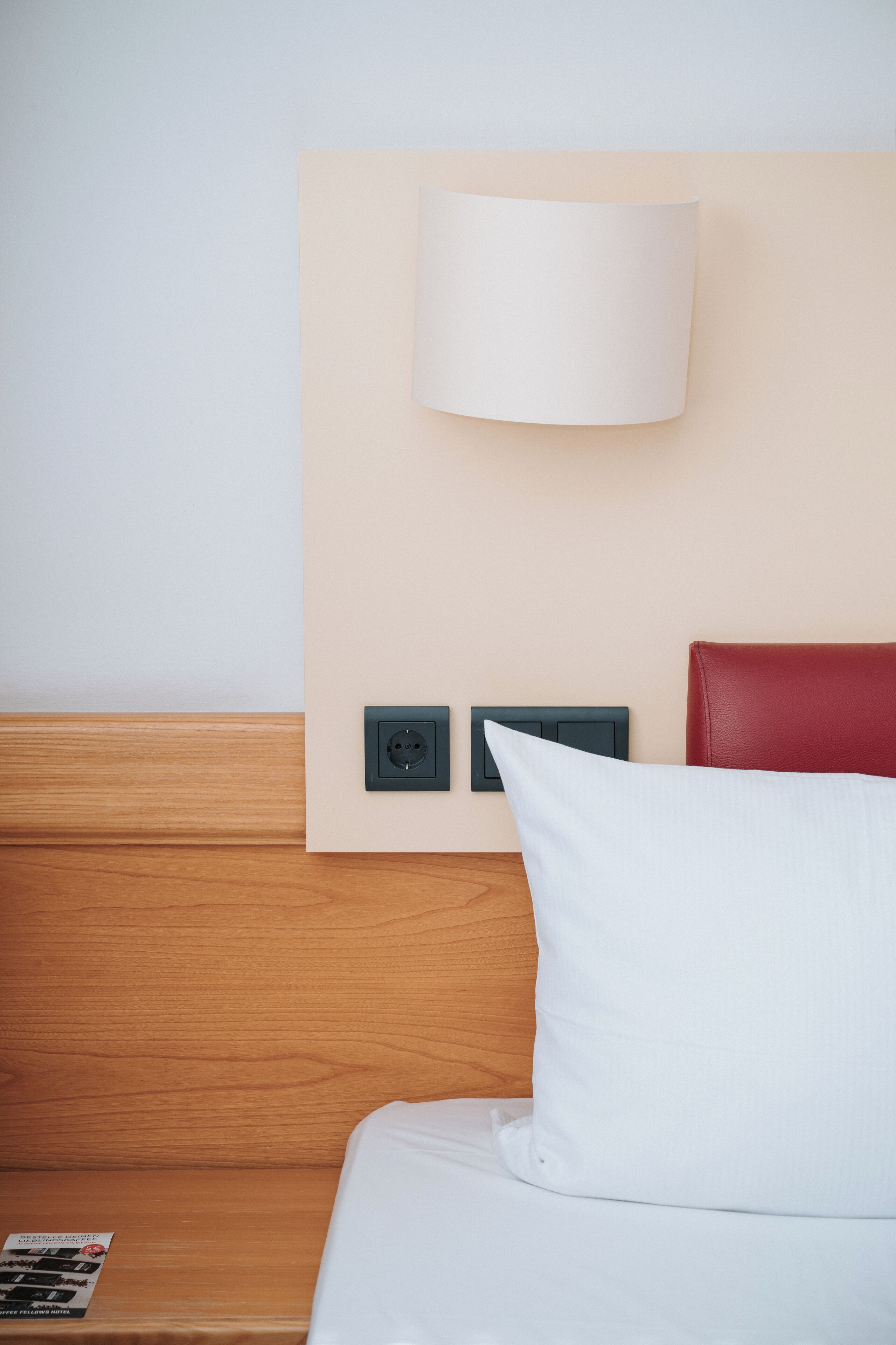 A hotel room with a wooden headboard, a white pillow, a wall-mounted lamp, and a power outlet.