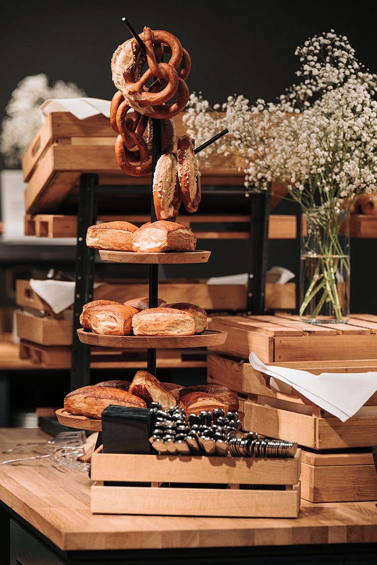 A wooden buffet table with various breads and pastries, including pretzels and baguettes, arranged on tiered wooden stands.