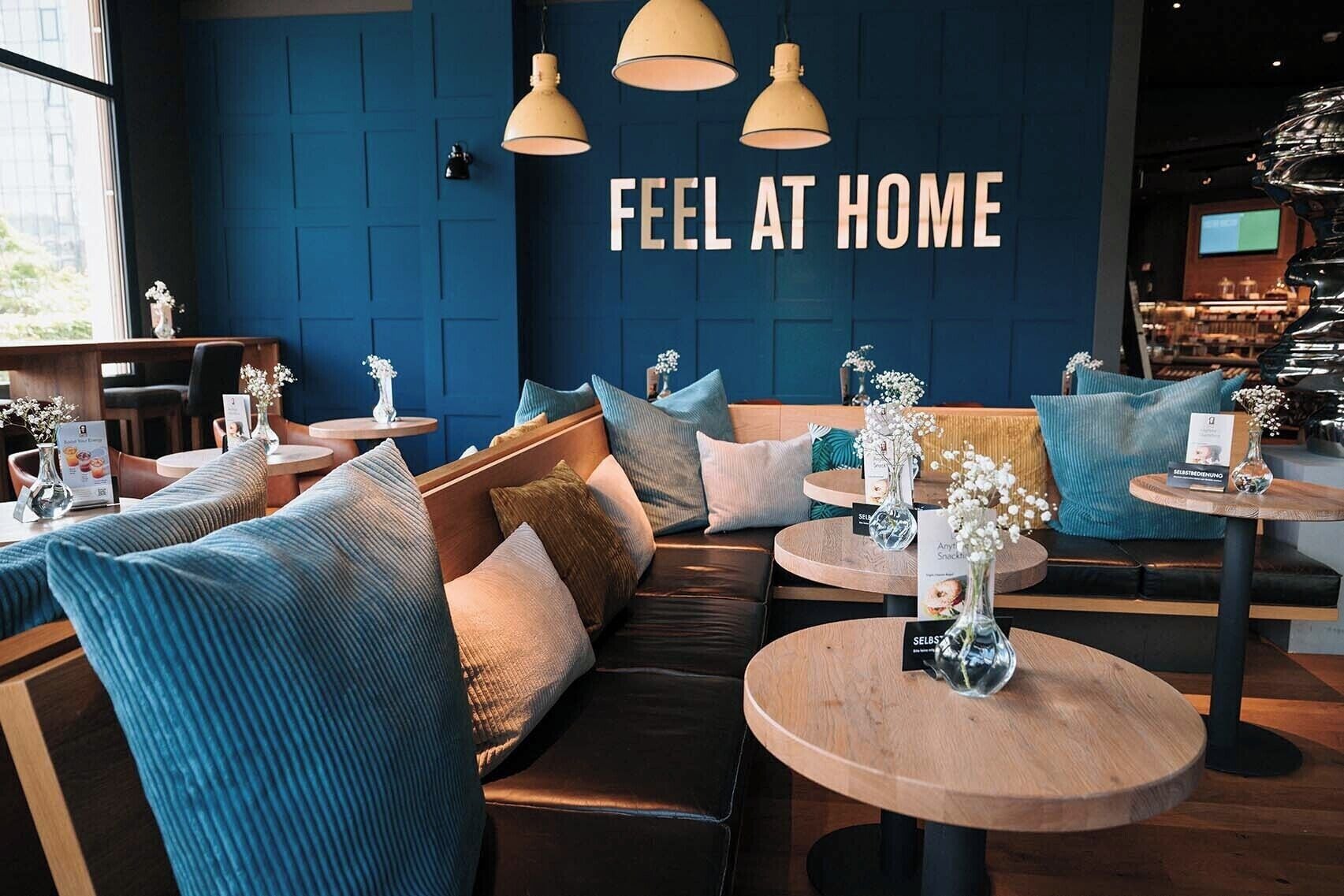 Un espace salon accueillant avec des canapés et des tables rondes, un mur bleu orné d’une inscription indiquant « VOUS ÊTES COMME CHEZ VOUS », et une vue sur la rue.