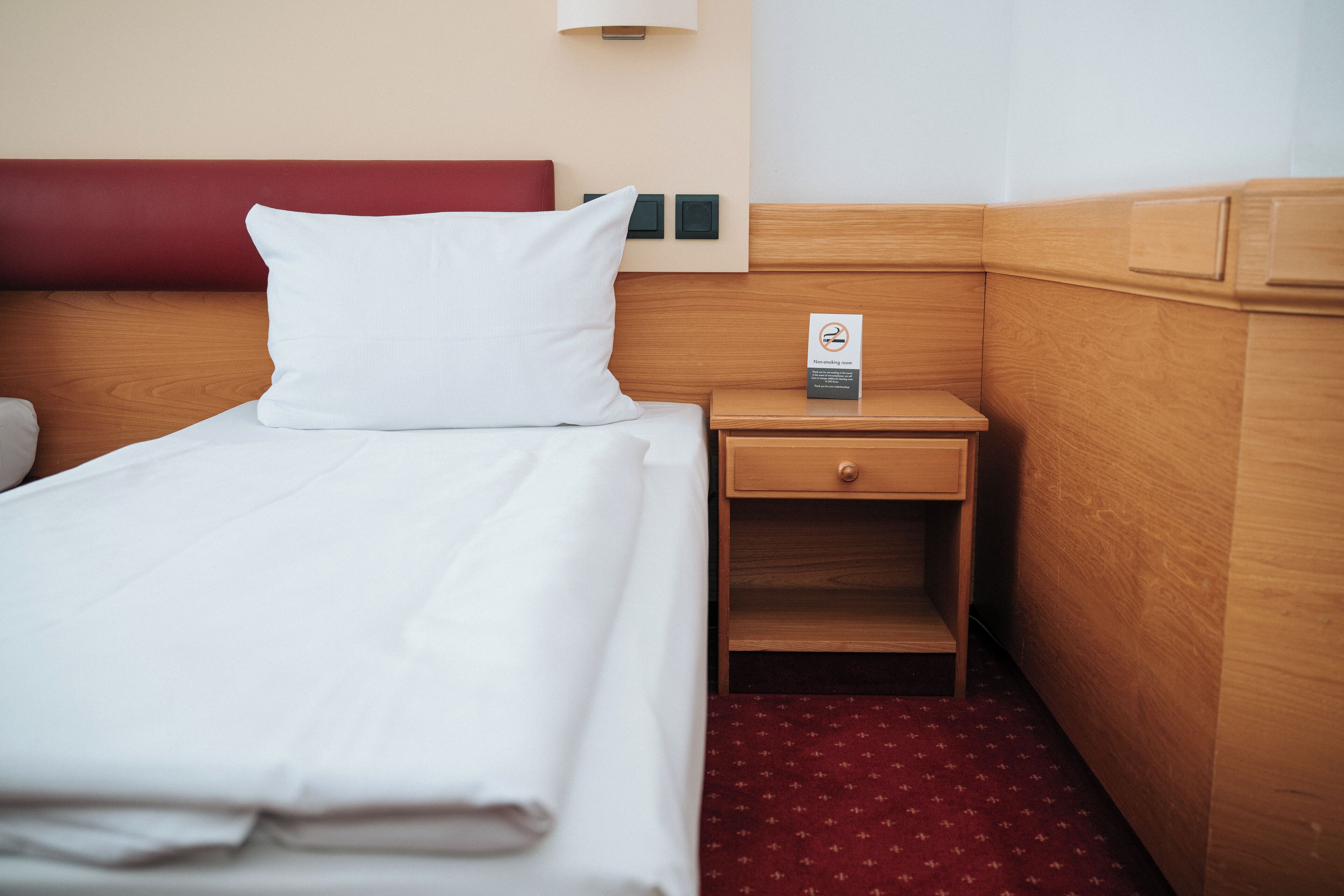 Ein Hotelzimmer mit einem ordentlich bezogenen Bett, einem Nachttisch aus Holz mit einer Lampe und einem roten Teppich mit Muster.