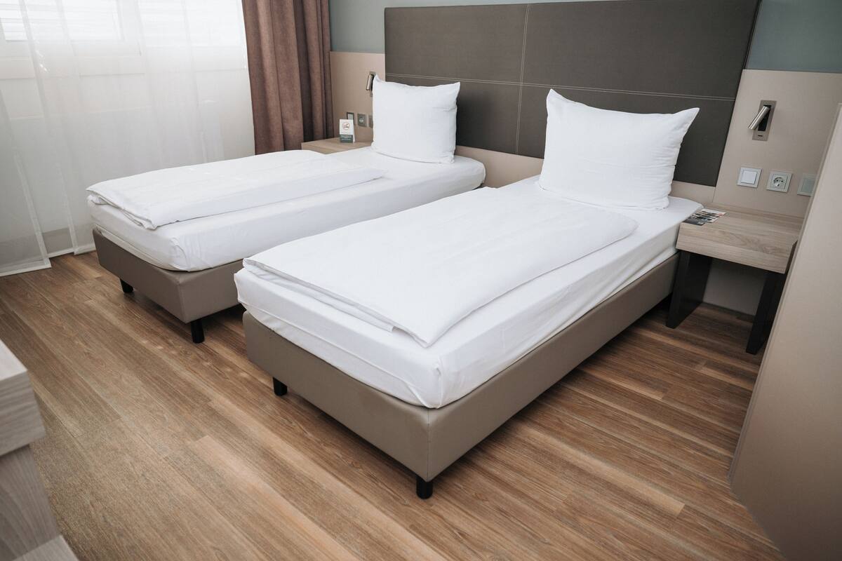 A hotel room with two single beds, wooden flooring, a nightstand with a lamp, and a window with curtains.