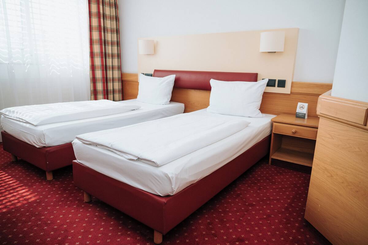 A hotel room with two single beds, a wooden nightstand, a red carpet, and a window with sheer curtains.