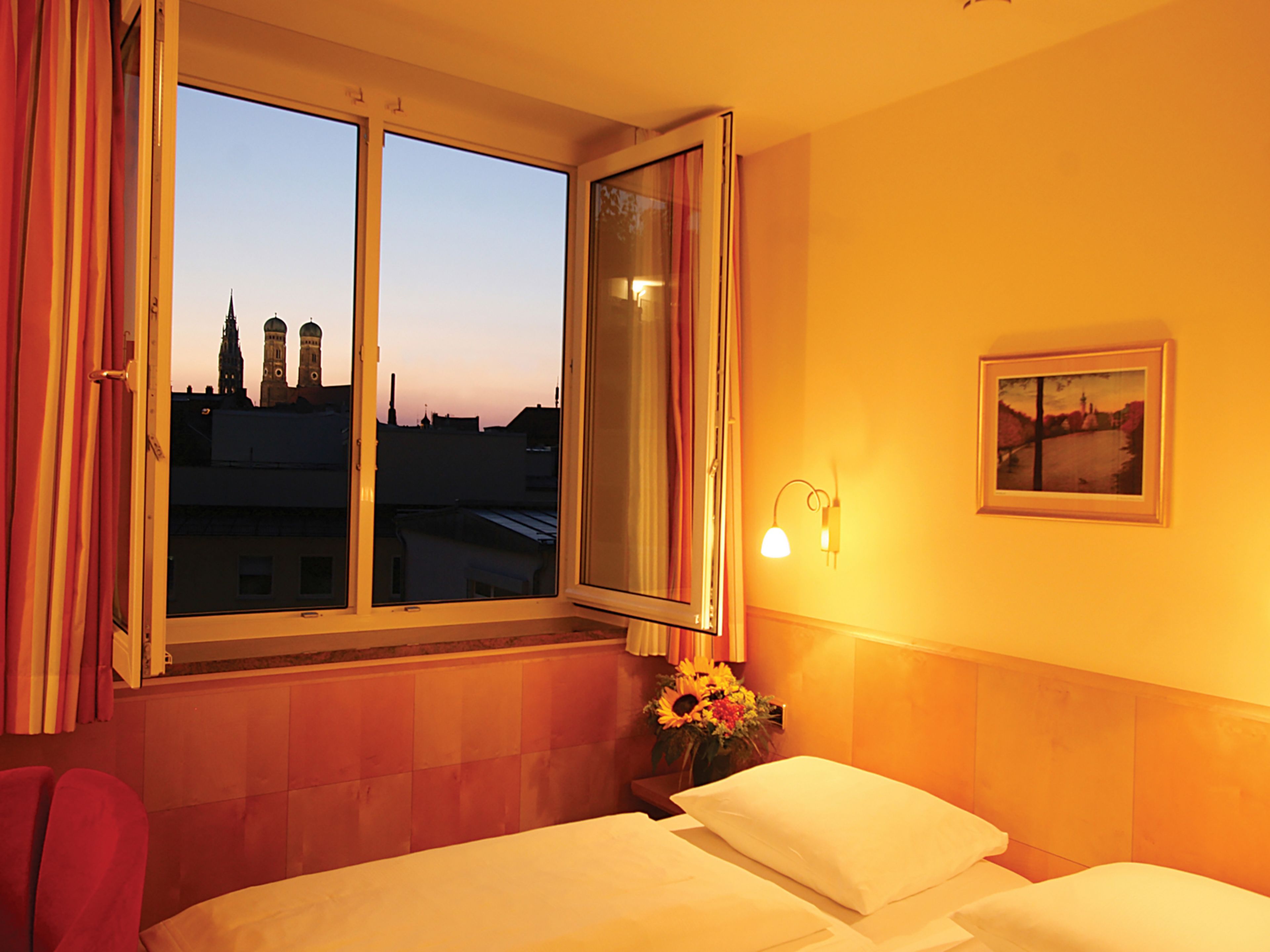 A hotel room with a bed, a nightstand with a lamp, a window with curtains, a painting on the wall, and a view of buildings outside.