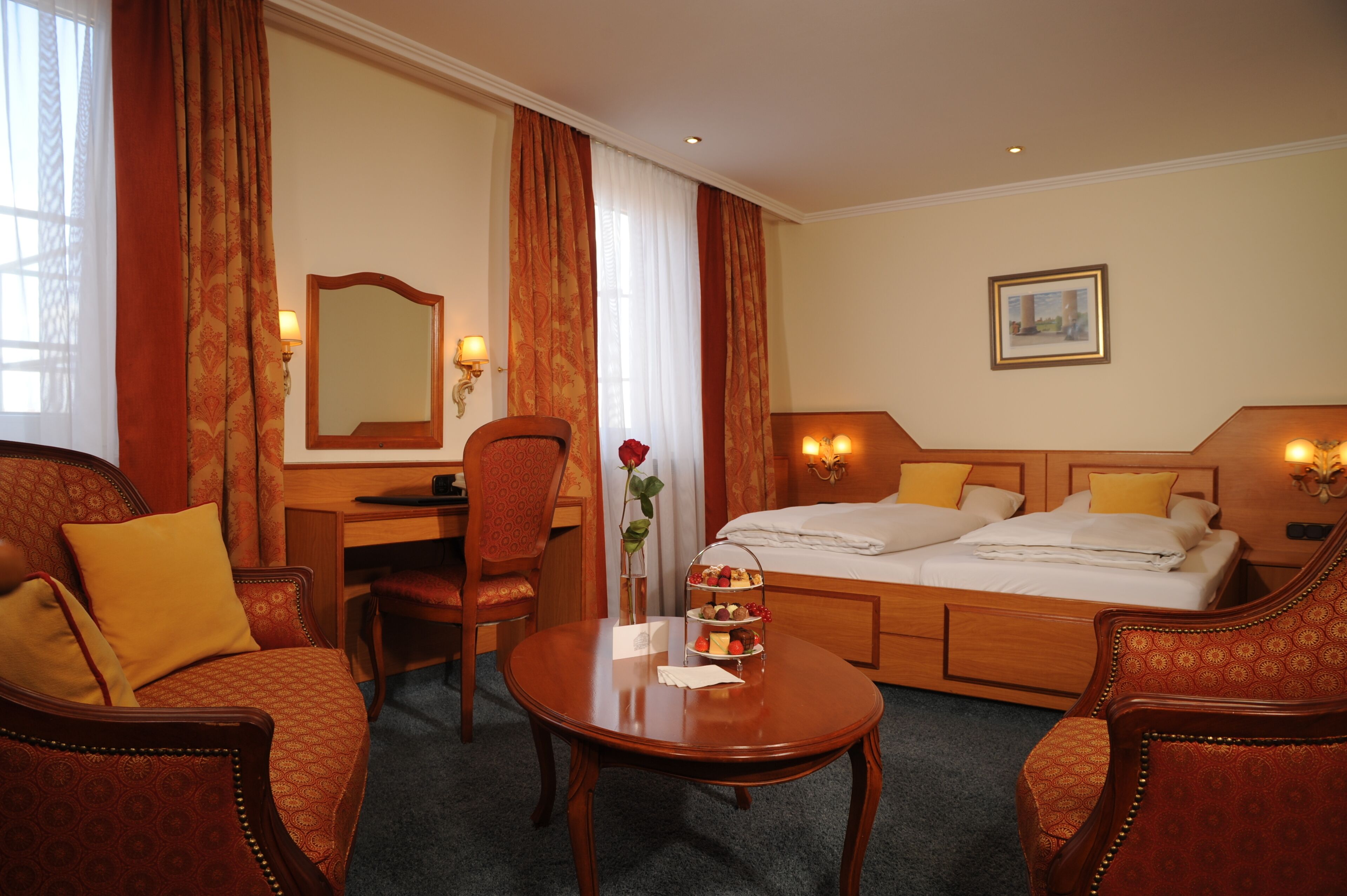 A hotel room with a bed, two chairs, a desk, a small table with a vase and a tray of fruit, and a painting on the wall.