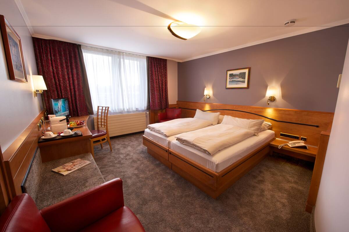 Habitación de hotel con cama doble, escritorio con silla, mesita de noche con lámpara, televisión y ventana con cortinas.