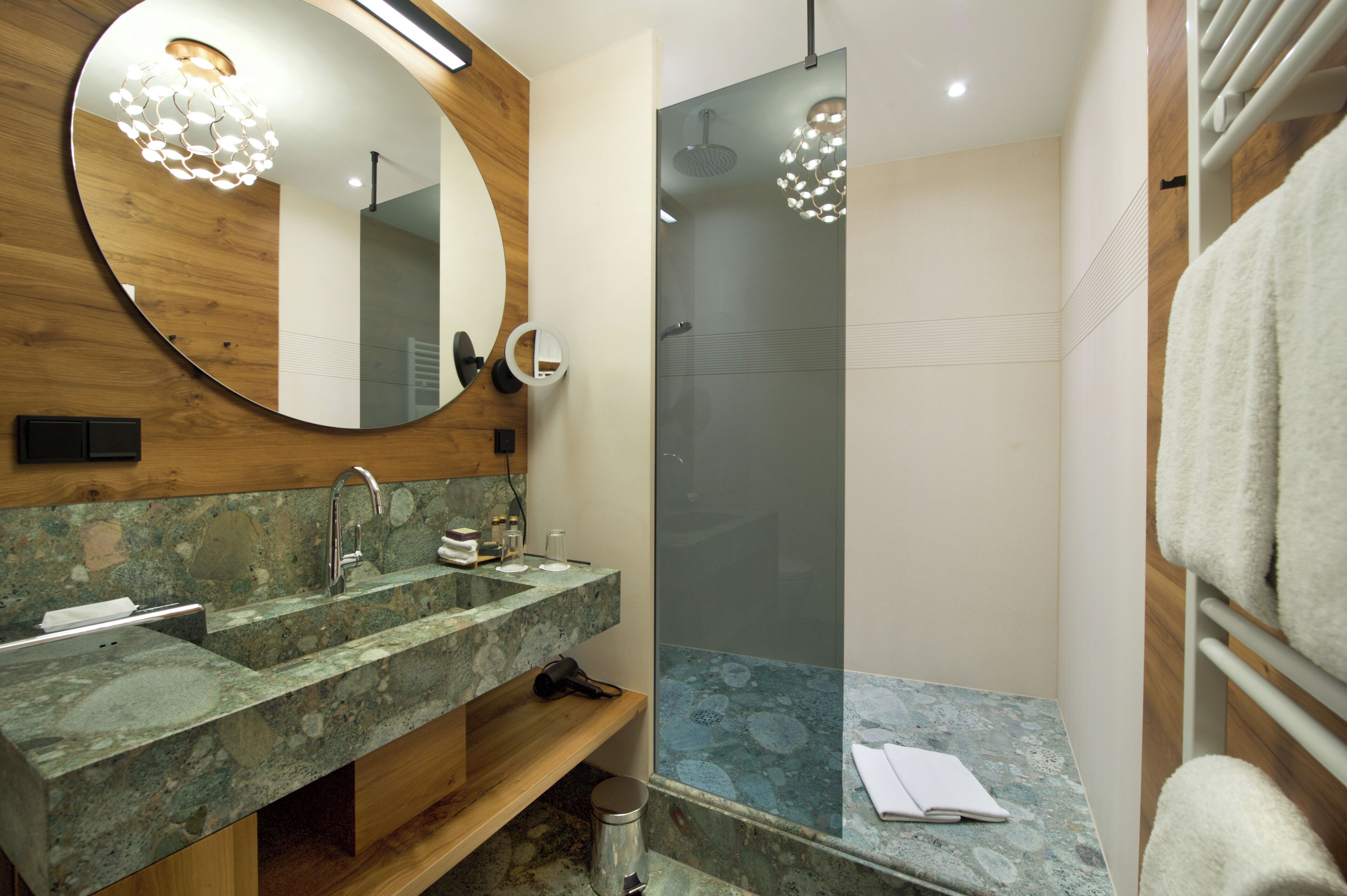 A modern bathroom with a wooden vanity, a glass shower enclosure, and a round mirror.