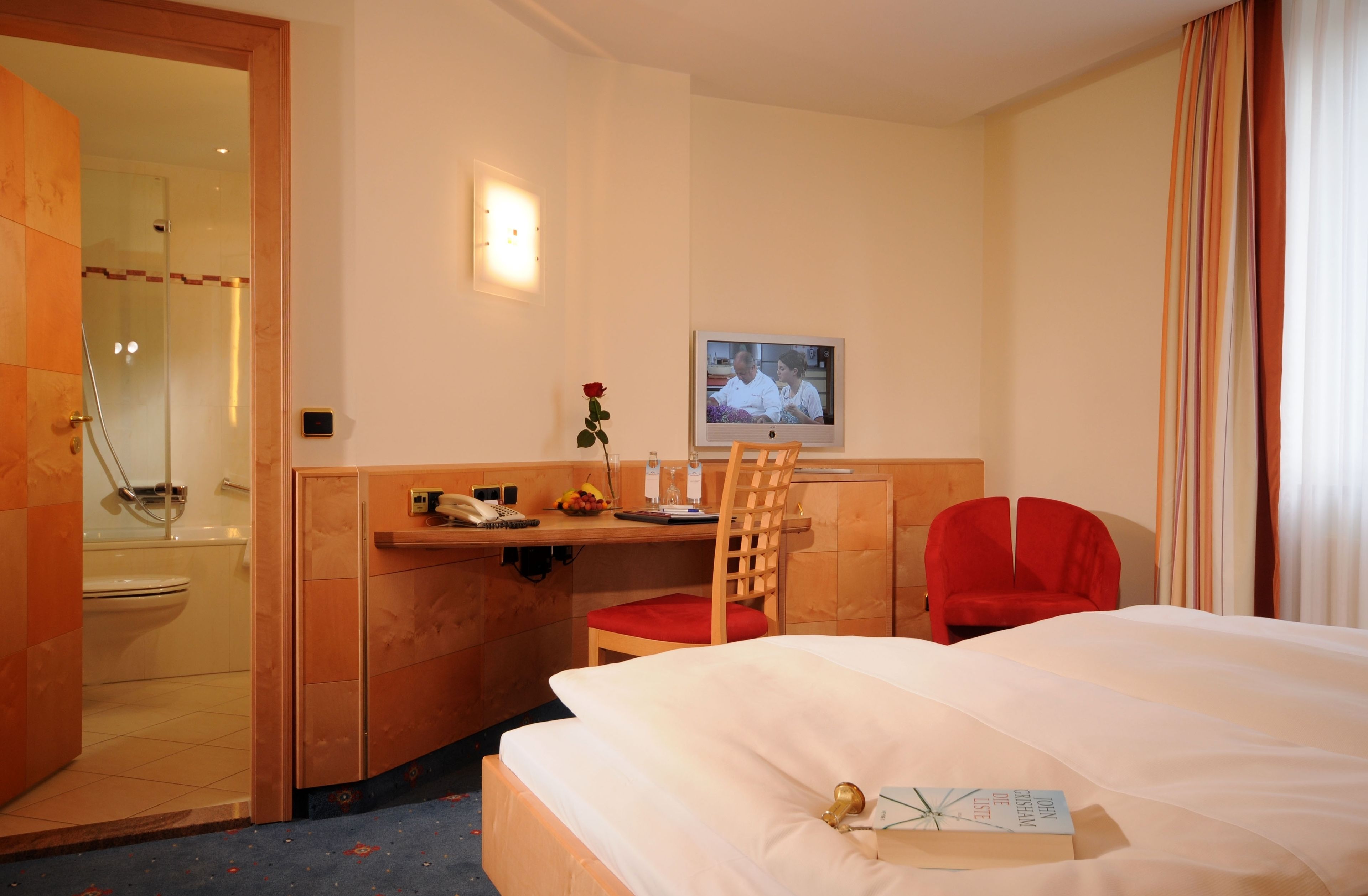 Quarto de hotel com cama, secretária com cadeira, chuveiro e televisão na parede.