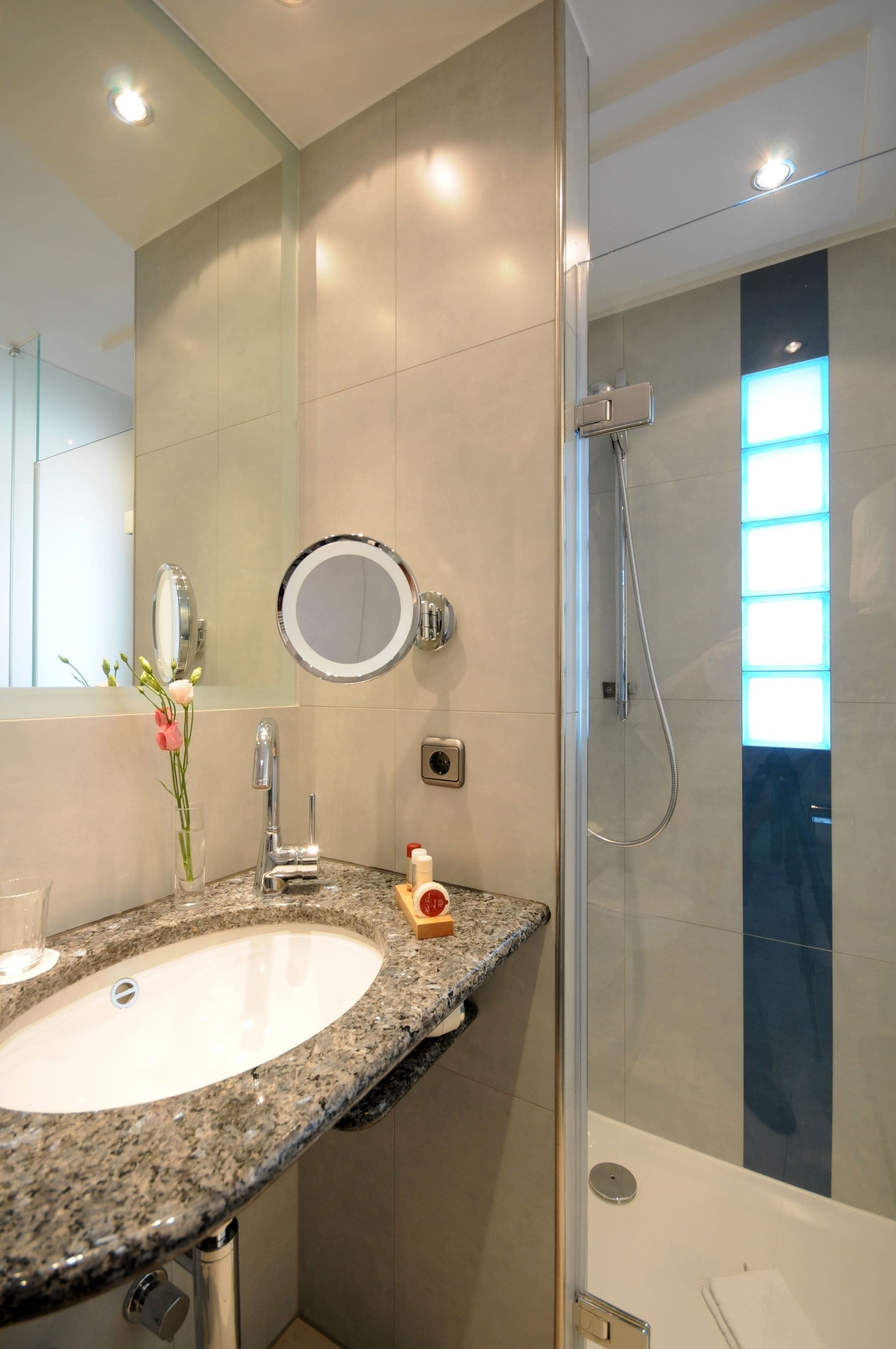 Une salle de bain moderne avec un lavabo surmonté d’un plan de travail en granit, un miroir rond et une douche à parois de verre.
