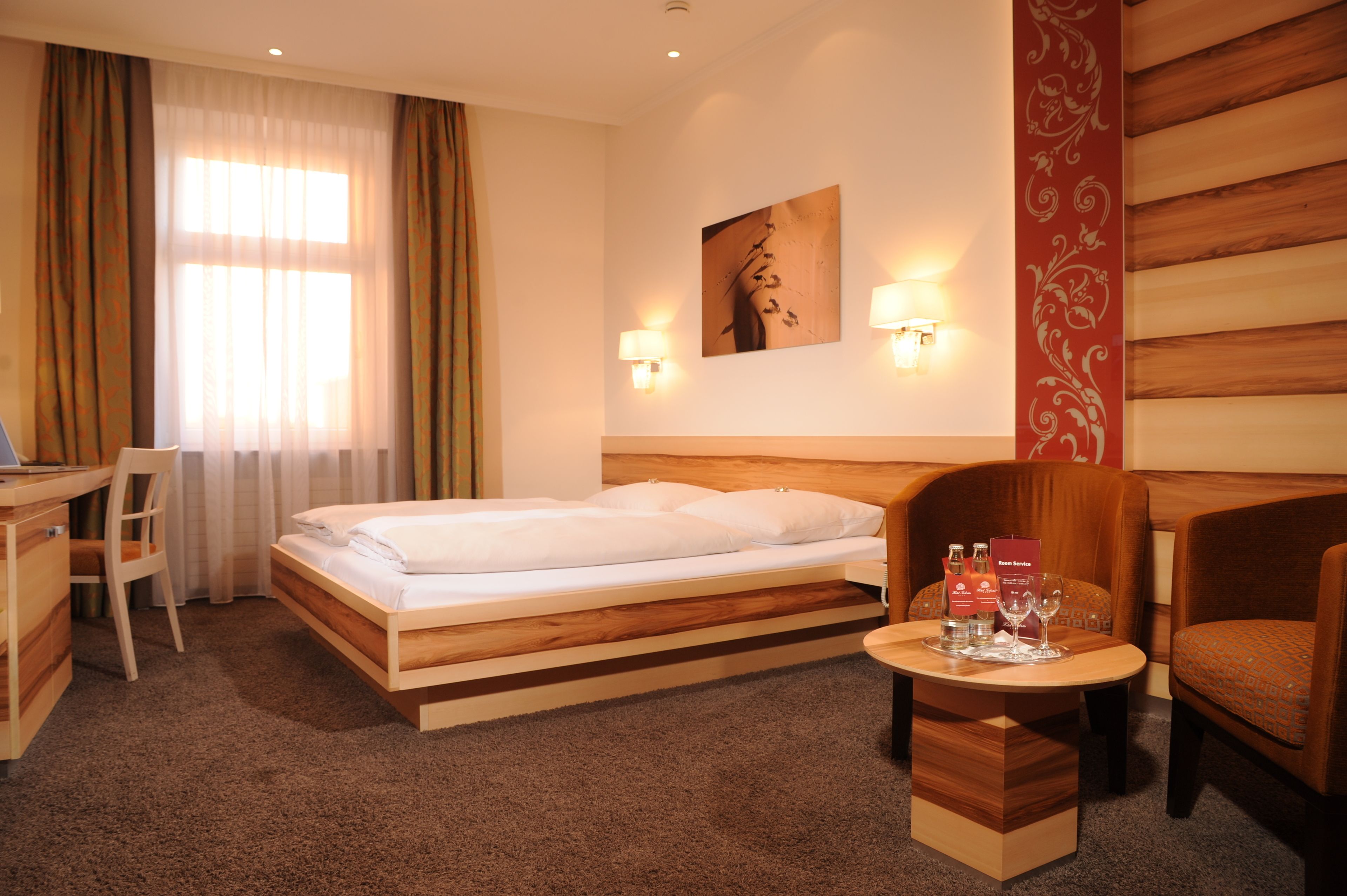Habitación de hotel con una cama, un escritorio, una silla, una mesita con una botella y un vaso, y una pared con un panel decorativo.