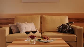 Um sofá bege com almofadas estampadas, um tabuleiro de vidro com petiscos e duas taças de vinho tinto.