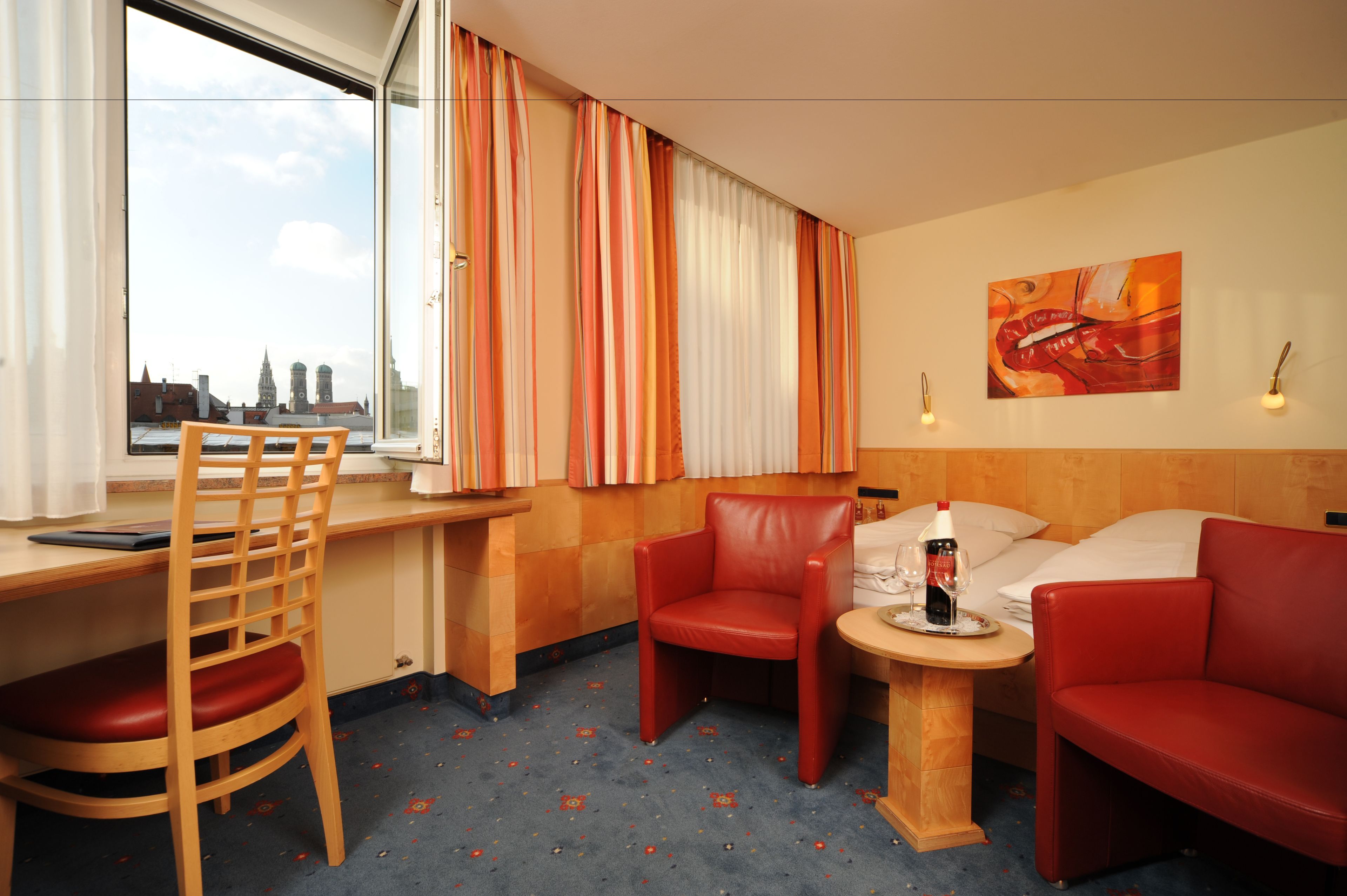 Et hotelværelse med udsigt over byen, et skrivebord, en stol, en sofa og et lille bord med en flaske og glas.