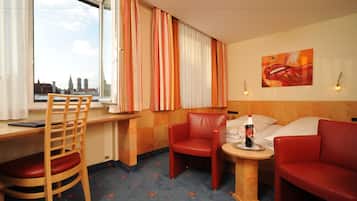 A hotel room with a city view, a desk, a chair, a sofa, and a small table with a bottle and glasses.