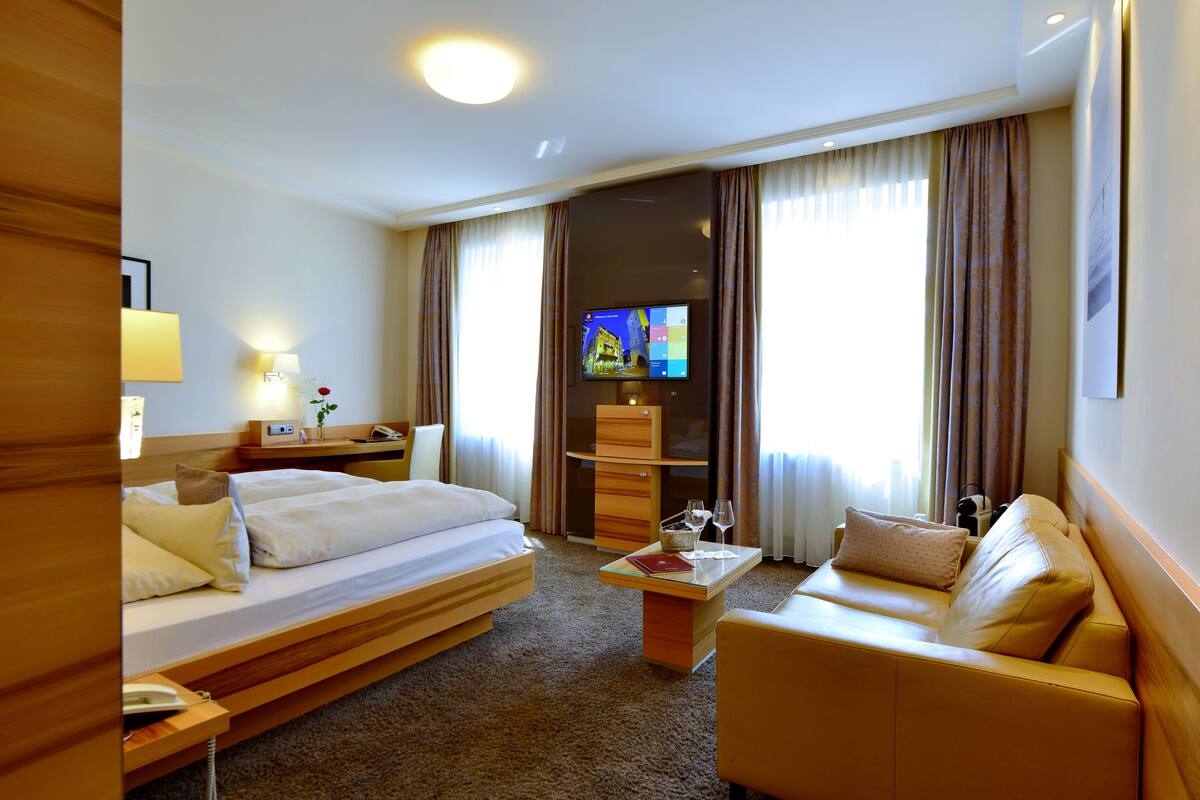 Una habitación de hotel con cama, sofá, televisor, escritorio y ventana con cortinas.