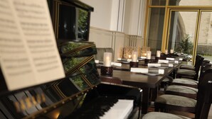 Huoneessa on piano, jonka päällä on nuotteja, ruokapöytä, jossa on kynttilöitä, sekä tuoleja. Huoneessa on suuret ikkunat.