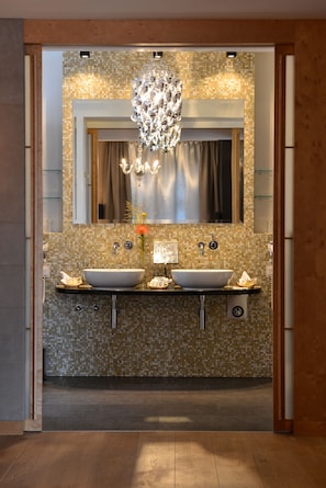 A modern bathroom with a mosaic tile backsplash, two sinks, a large mirror, and a decorative chandelier.