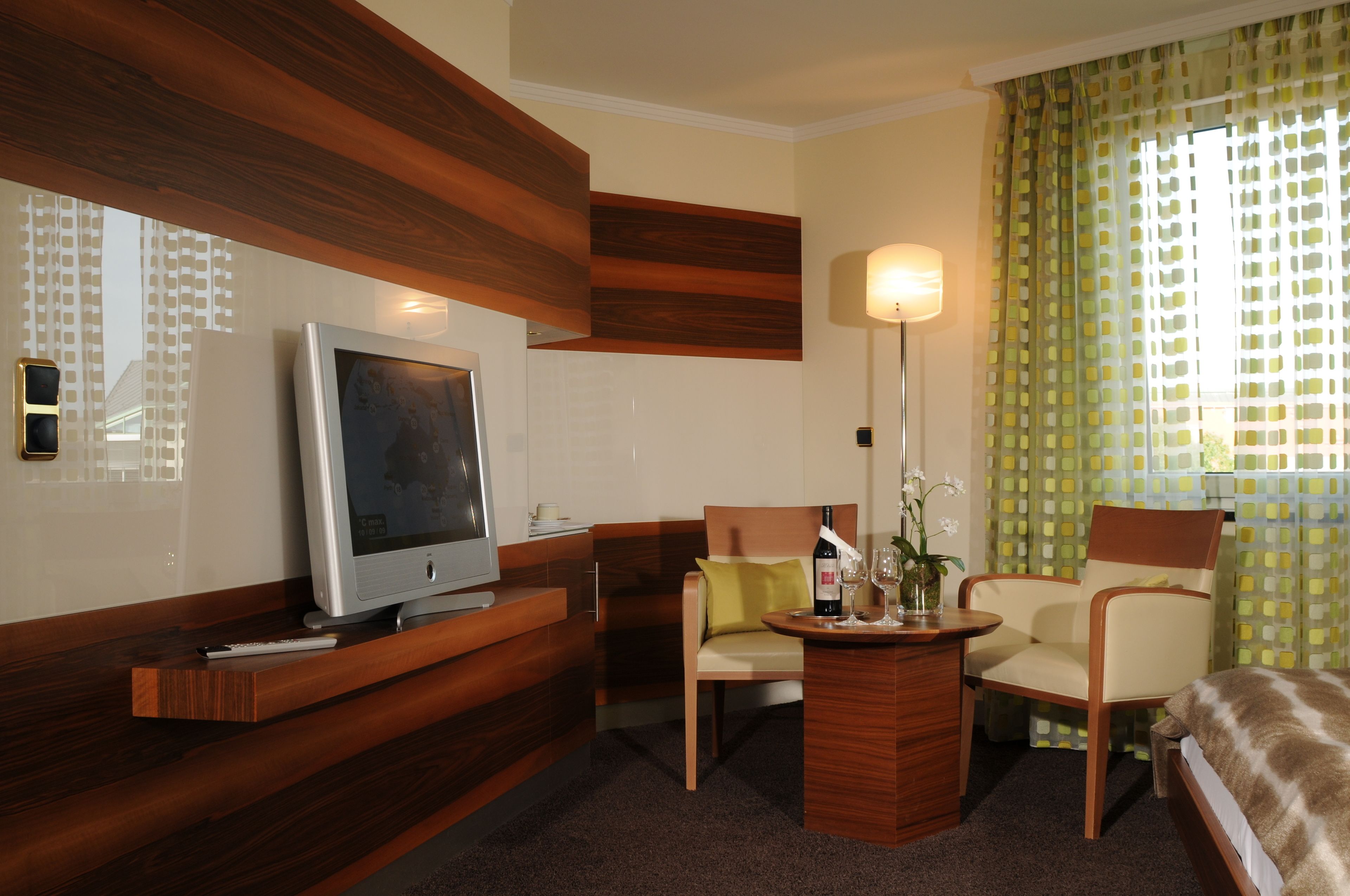 A hotel room with a television, a sofa, a chair, a small table with a bottle and glasses, and a window with patterned curtains.