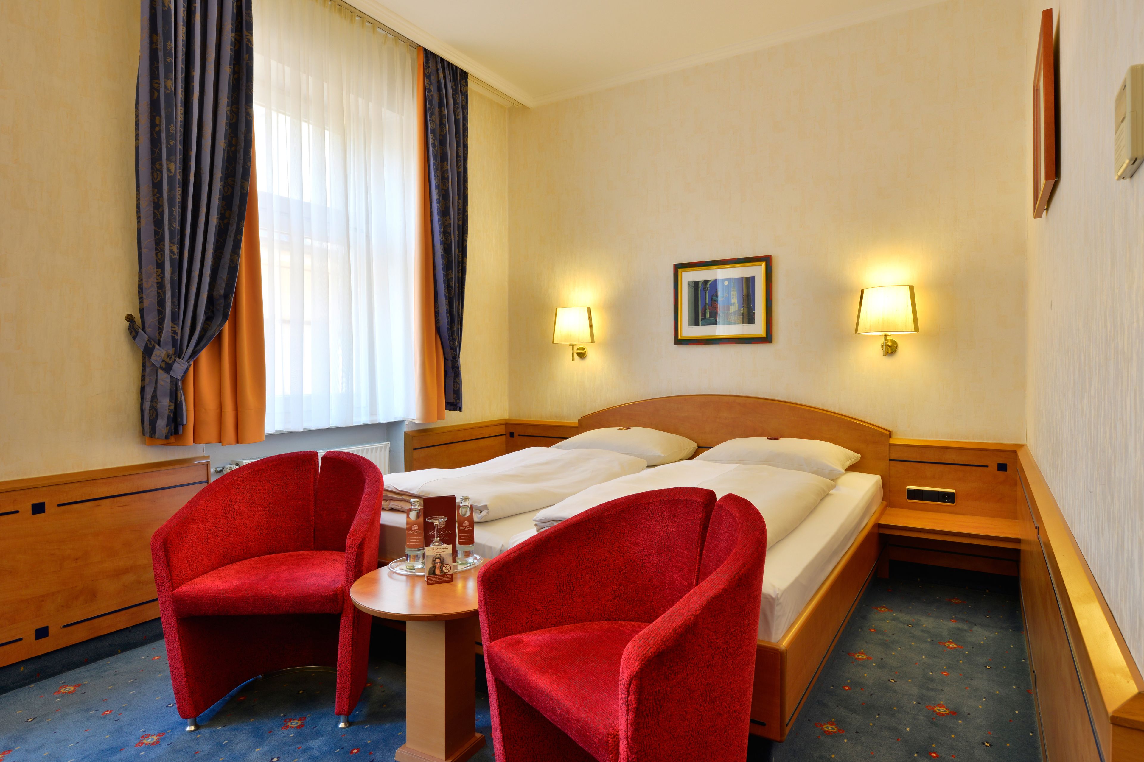 Ein Hotelzimmer mit einem Bett, zwei roten Sesseln, einem kleinen Tisch und zwei Wandleuchten.