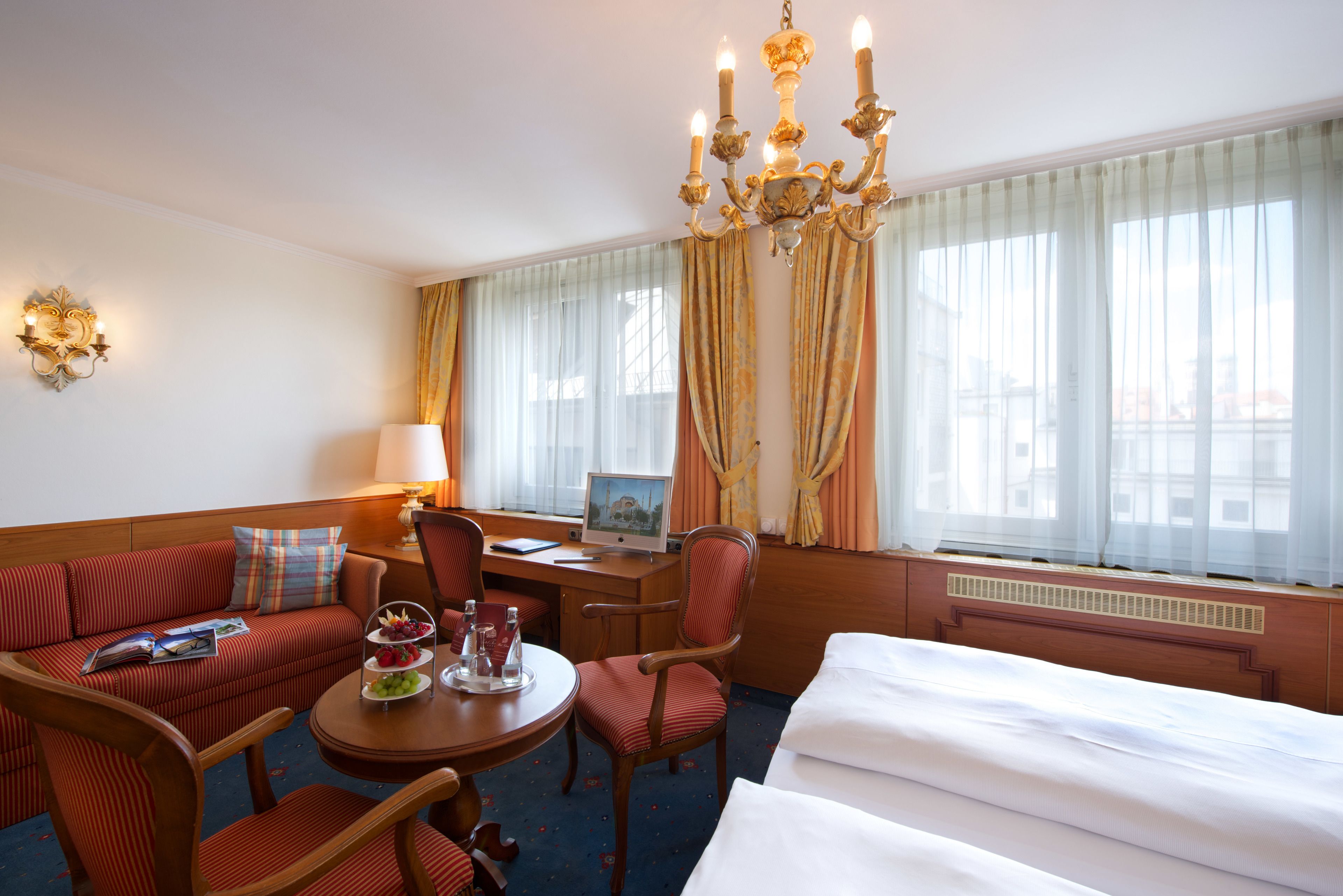 A hotel room with a bed, a sofa, a desk with a computer, a chair, and a chandelier.