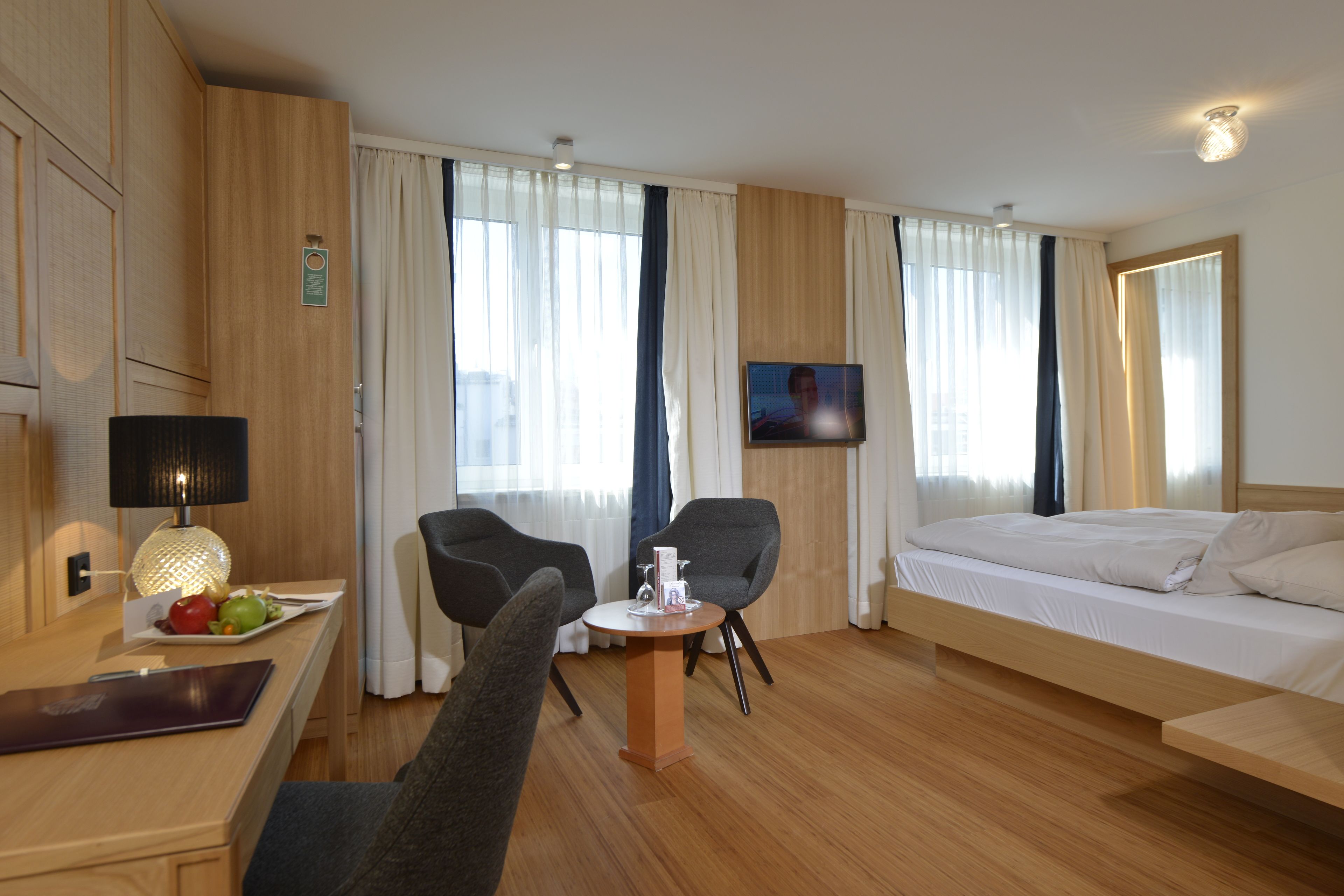 Habitación de hotel con una cama, dos sillas, una mesita, televisor y escritorio con lámpara y una bandeja de frutas.