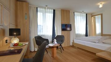 Habitación de hotel con una cama, dos sillas, una mesita, televisor y escritorio con lámpara y una bandeja de frutas.