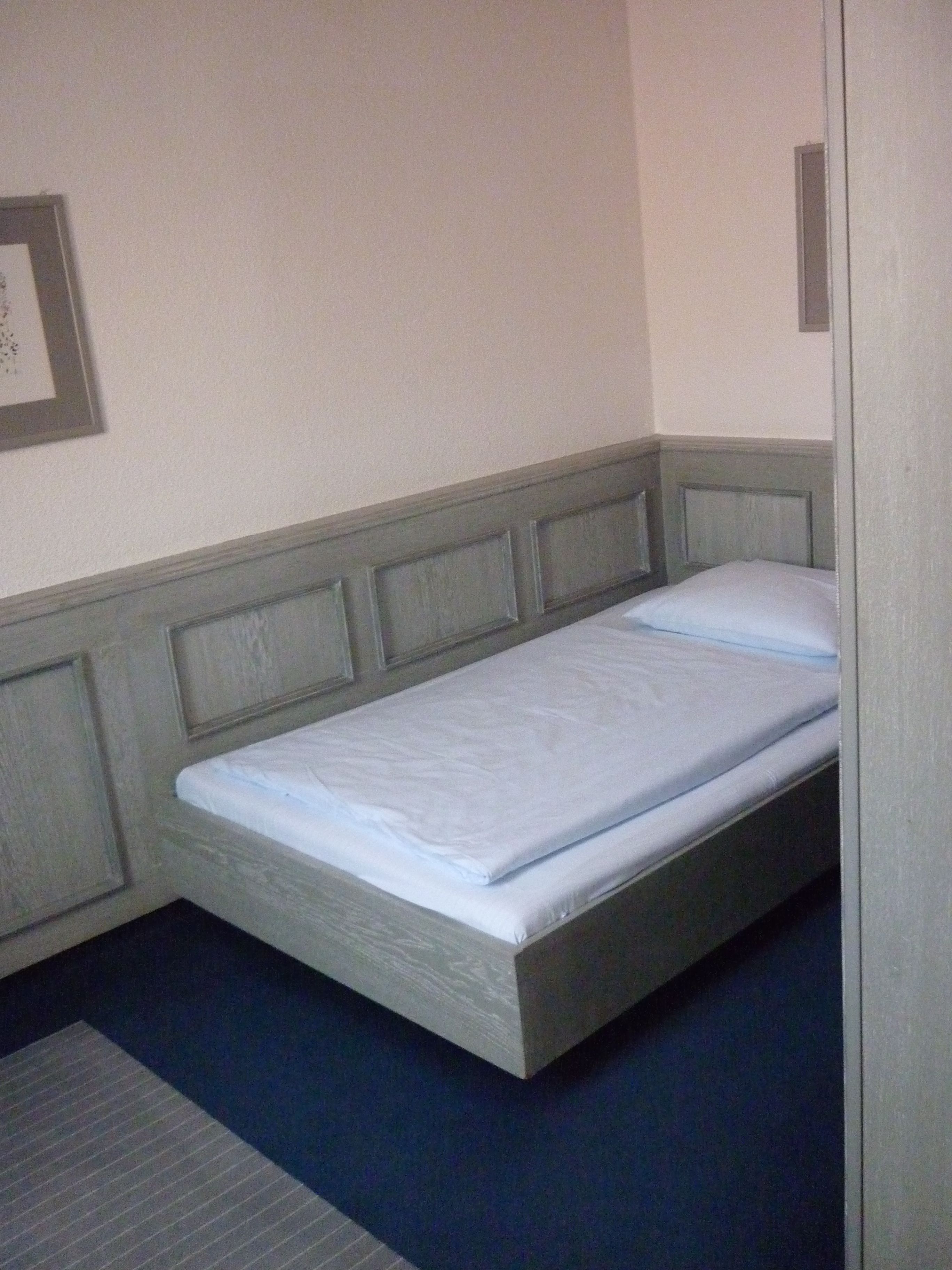 Una cama individual con cabecera de madera y colchón azul en una habitación con paredes lisas y suelo oscuro.