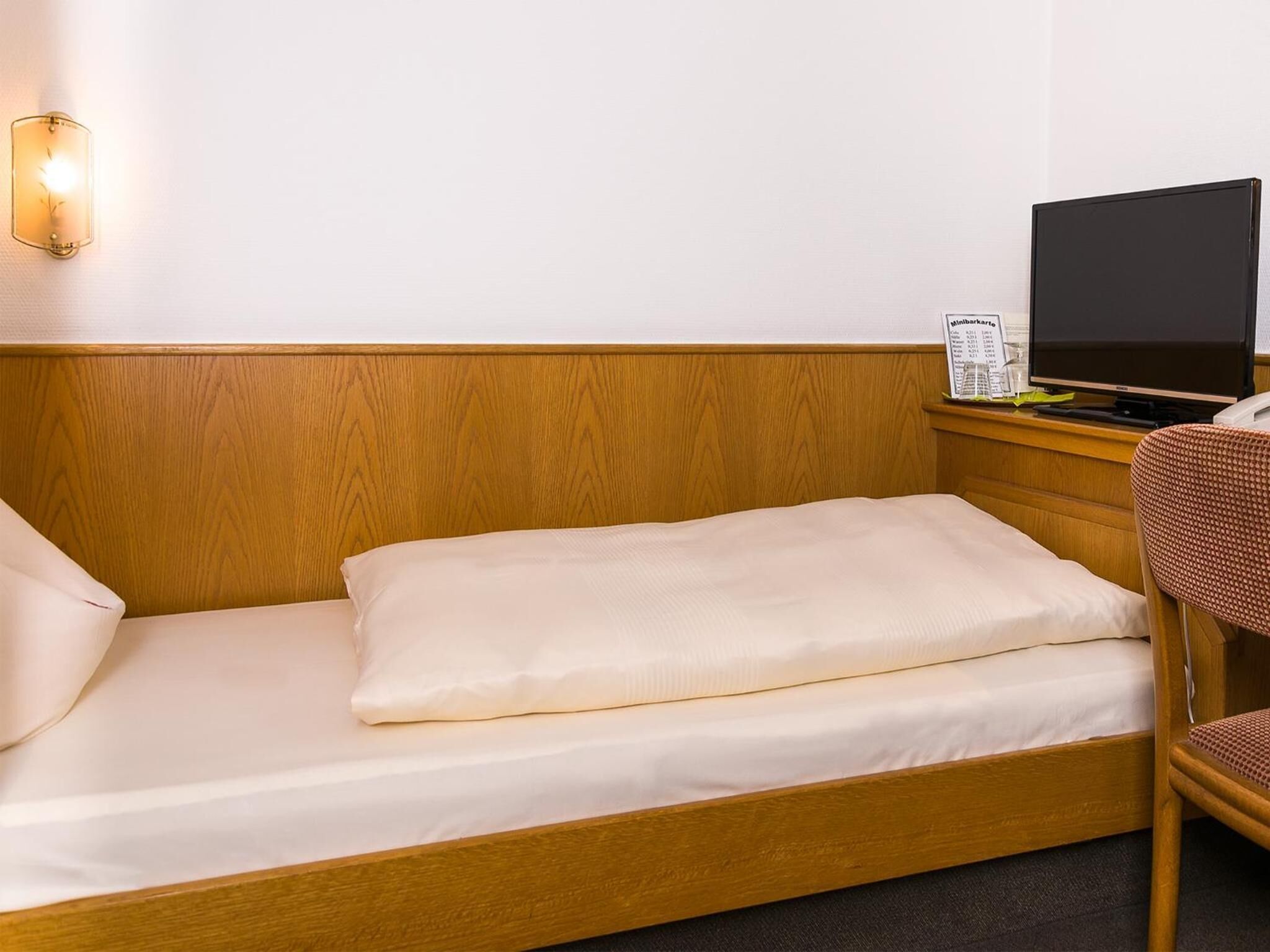 Una cama individual con cabecera de madera, una mesita de noche con televisor de pantalla plana y una silla con asiento de mimbre.
