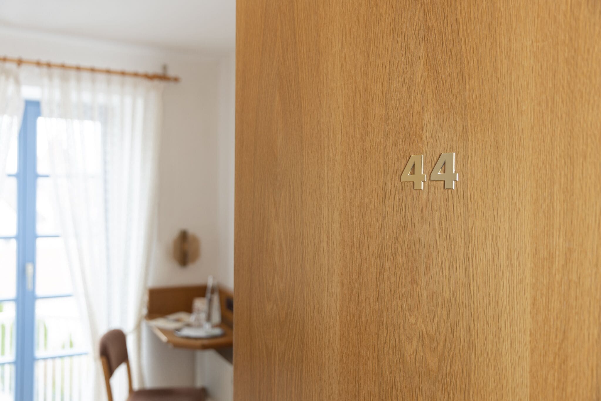 Una porta de fusta amb el número 44, una finestra amb cortines lleugeres, i un interior borrat amb una taula i una cadira.