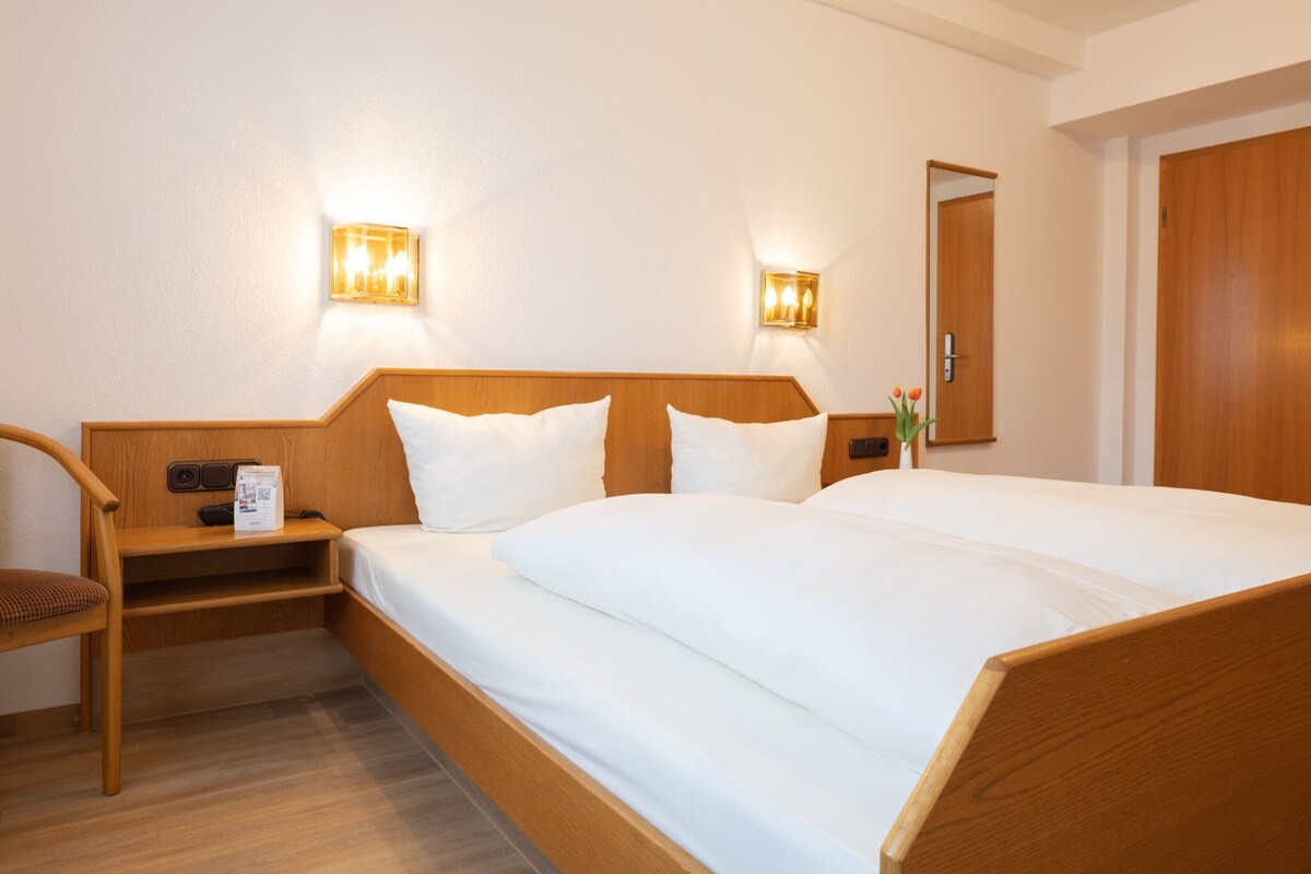 A hotel room with a wooden bed, two bedside tables, a chair, and two wall-mounted light fixtures.