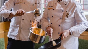 Two chefs in a kitchen, one holding a ladle and the other holding a pan.