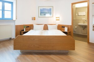 Una habitación de hotel moderna con una cama grande de madera, vistas a la ciudad, un baño con lavabo y espejo, y una pequeña mesa con un jarrón de flores.