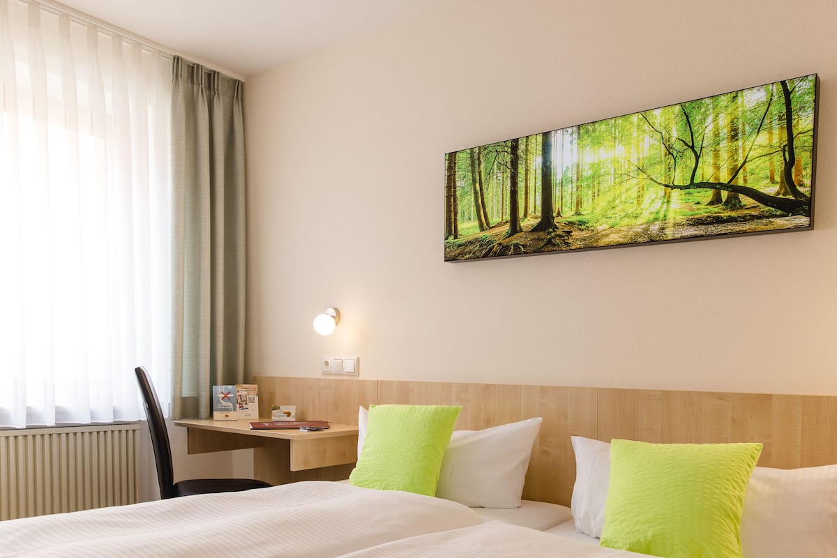 Habitación de hotel con cama, escritorio, silla, un cuadro de un bosque y ventana con cortinas.