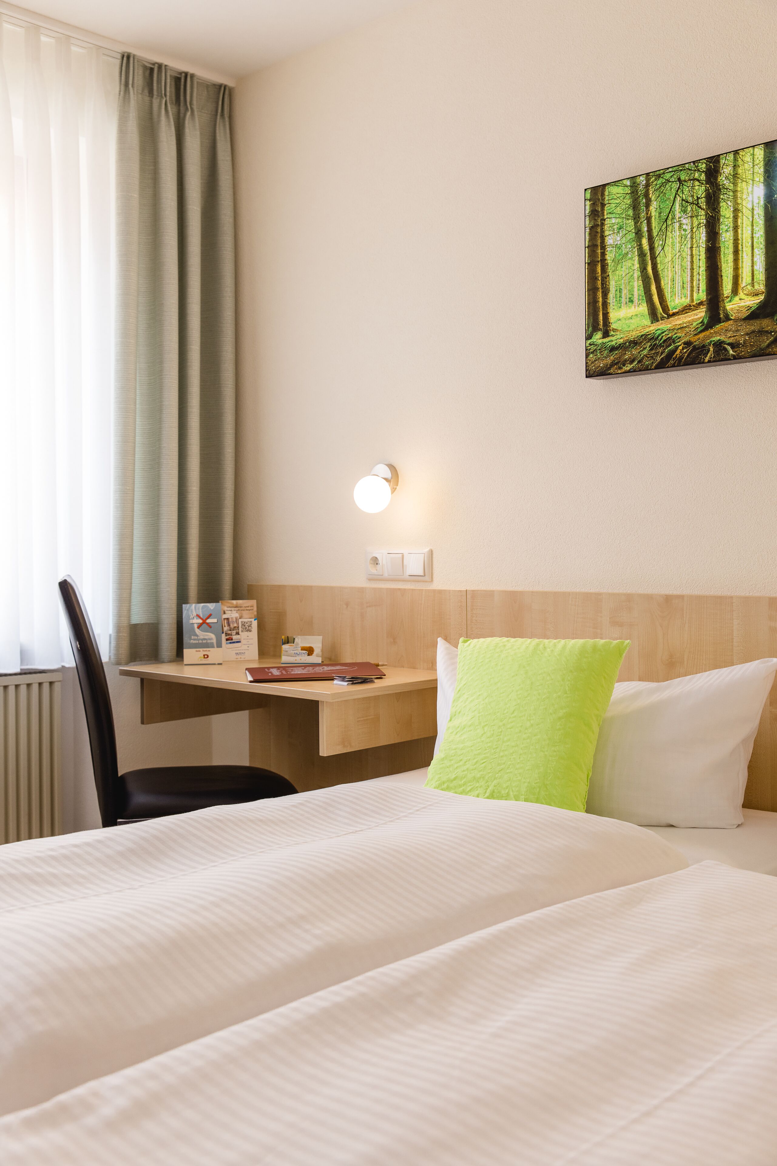 Habitación de hotel con cama, escritorio, silla, un cuadro de un bosque y ventana con cortinas.